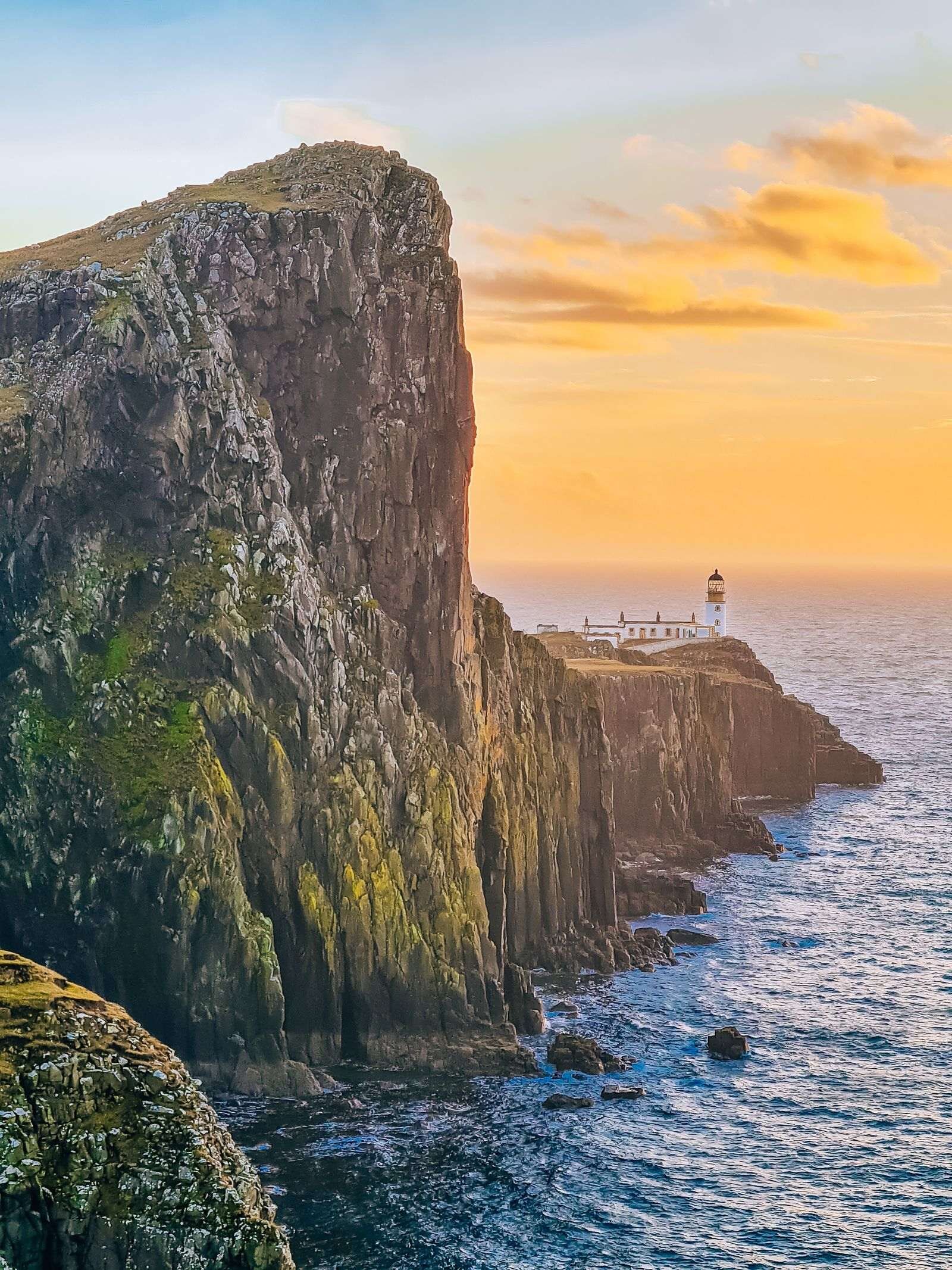 A tall rocky sea cliff which slopes down in the distance with a lghhouse on the end of the rocky peninsula. The sky is orange at sunset.