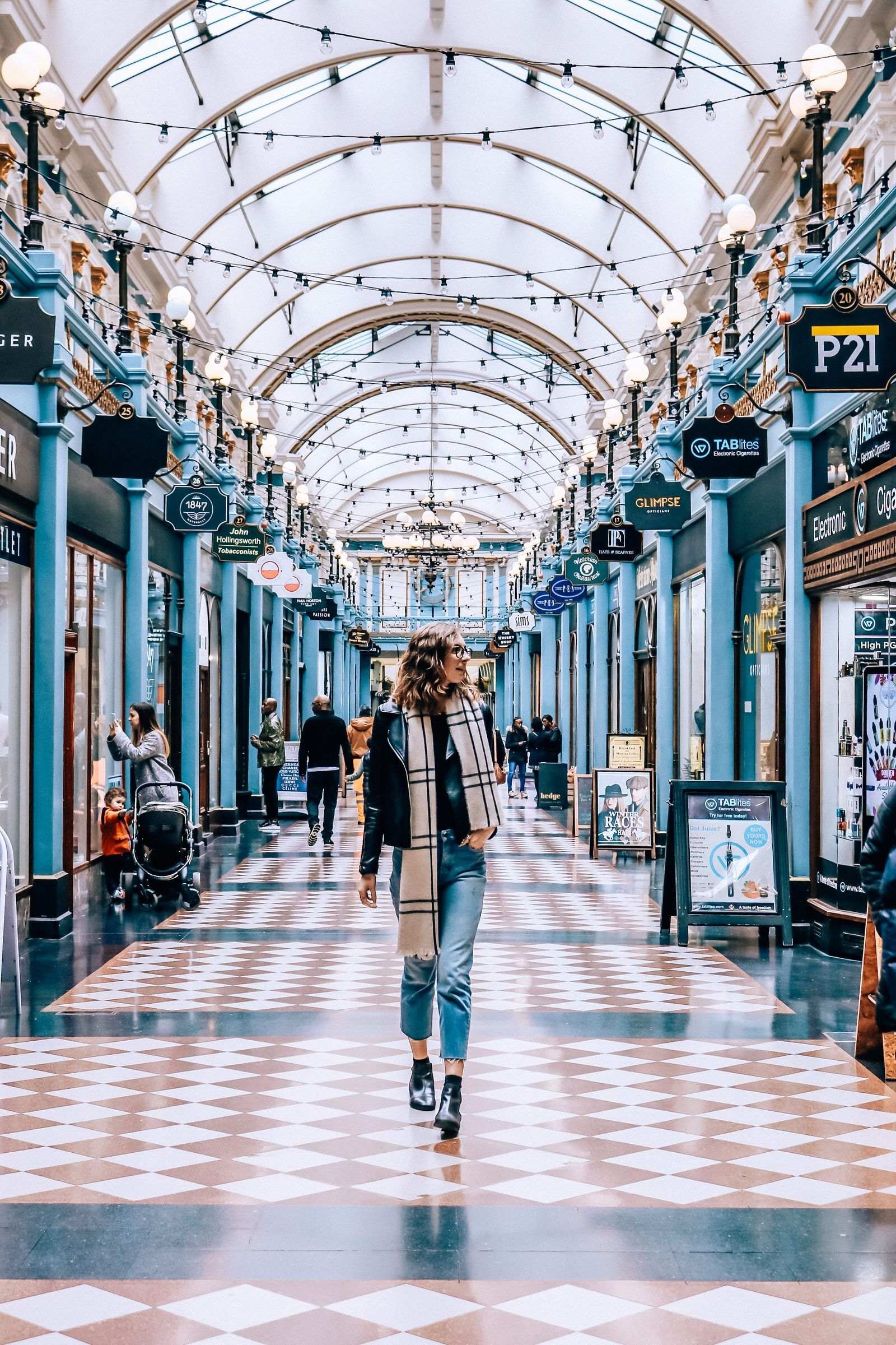 An indoor shopping arcade with blue pillars and diamond patterned floor, woman is walking down the centre of the arcade in jeans, boots and a black jacket