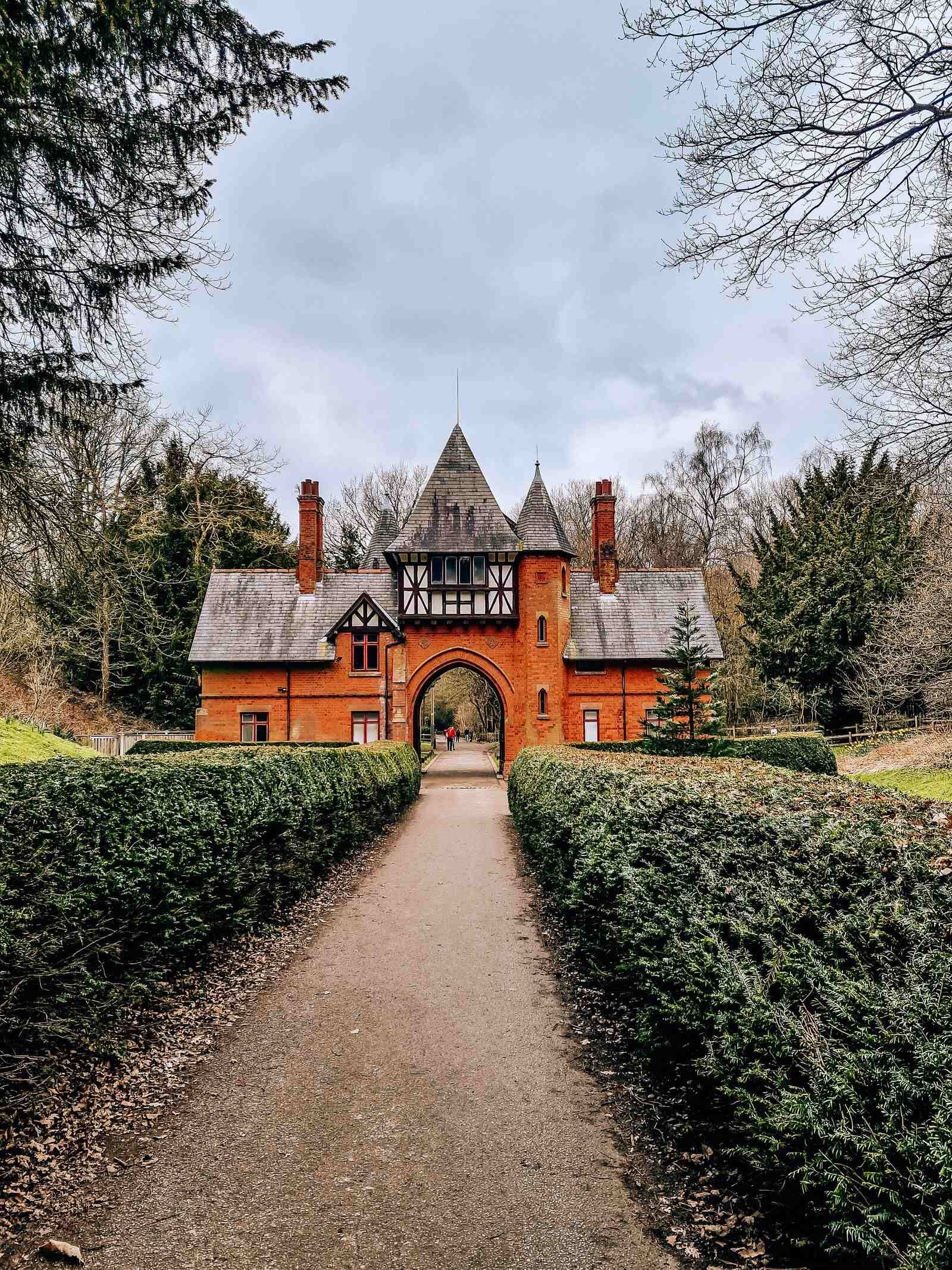 a red brick gatehouse with a timber framed roof and slate tiles. Path leading to the gae is lined with green hedges and there are trees behind the gatehouse