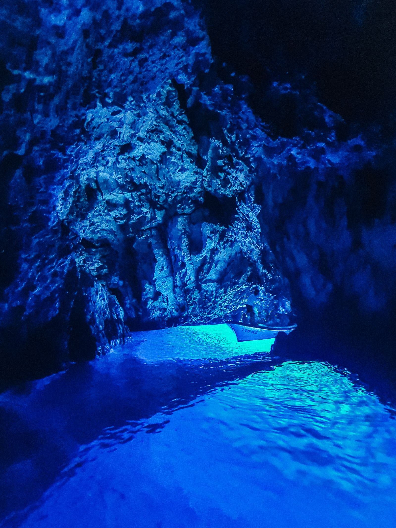 Inside a dark cave, the only light is blue light coming up from the water below. A small white boat can be seen further into the cave