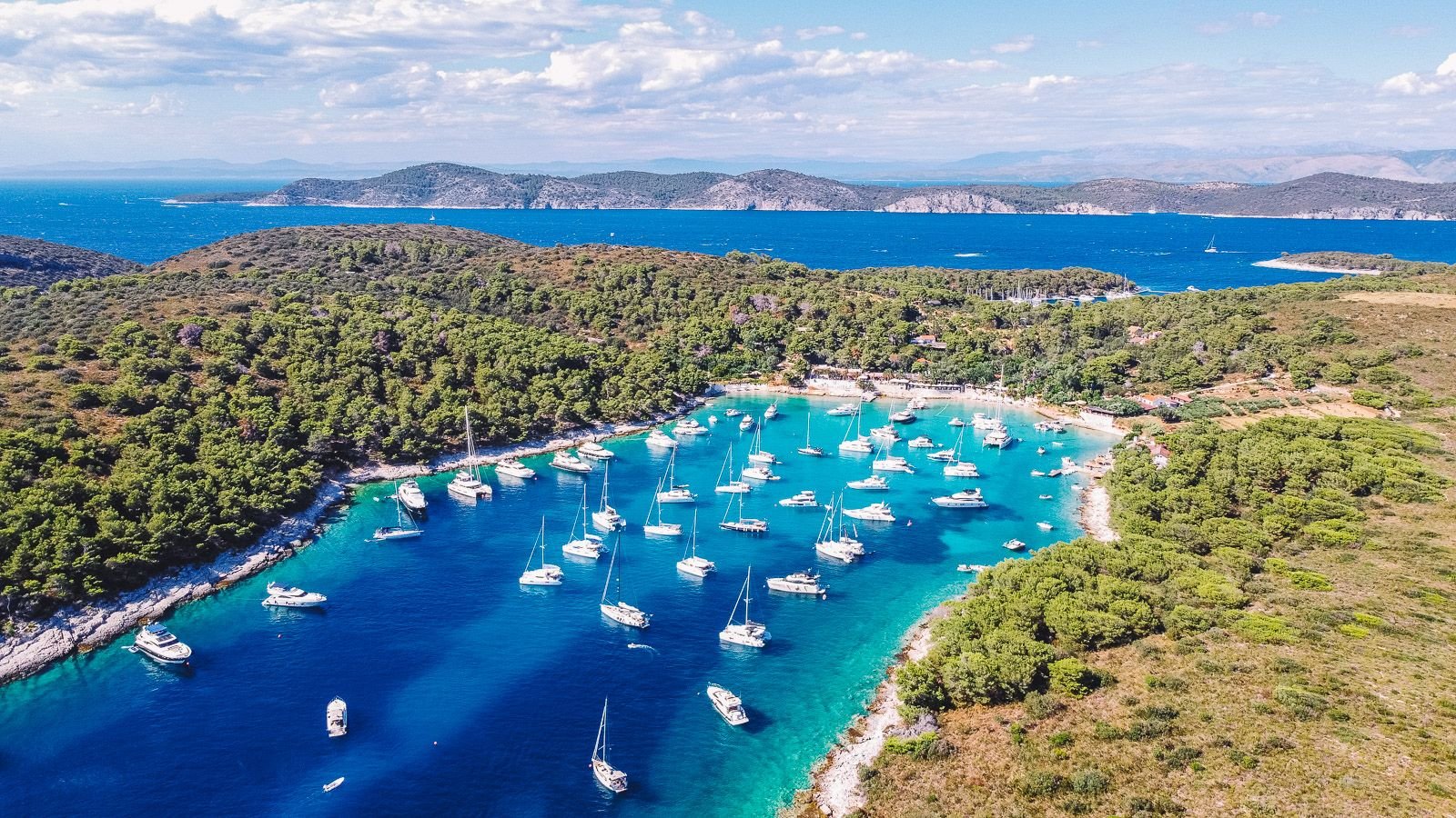 Drone shot of a long turquoise blue pay with many yachts and sailing boats anchored. The surroundings of the beach are lush green covered in trees and moutains and more islands are in the distance