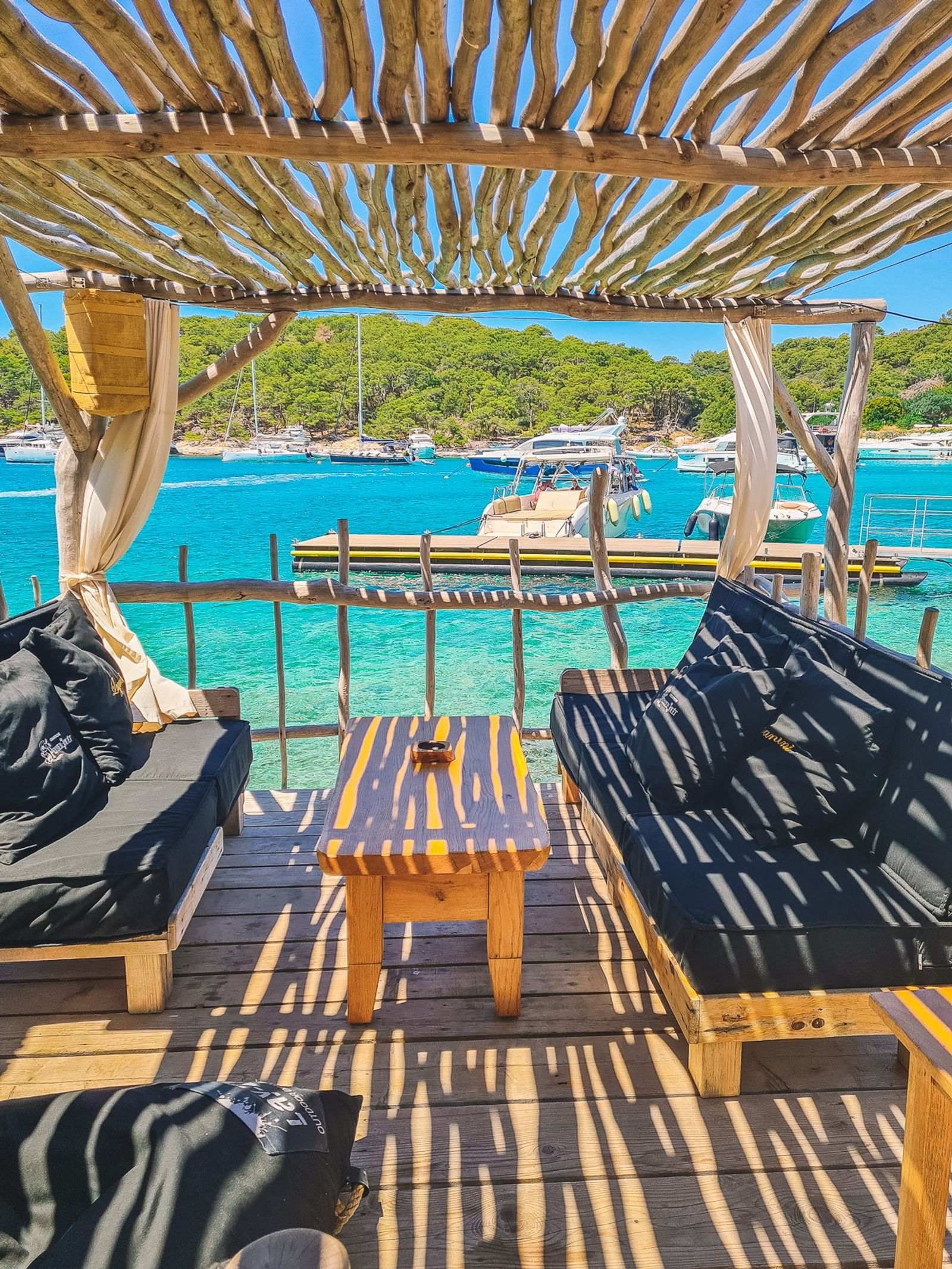 a beachside cabana made of driftwood with black sofas in the shade either side of a table. Right next to shallow turquoise water on Pakleni Islands in Croatia