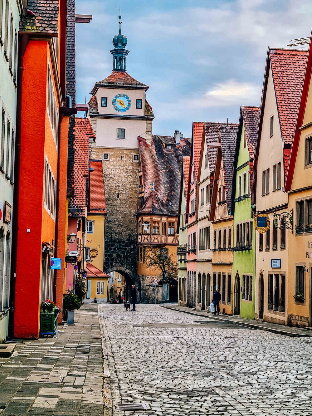  A cobbled street lined with colourful buildings on each side leading up to a stone walled tower with a clock near its top 