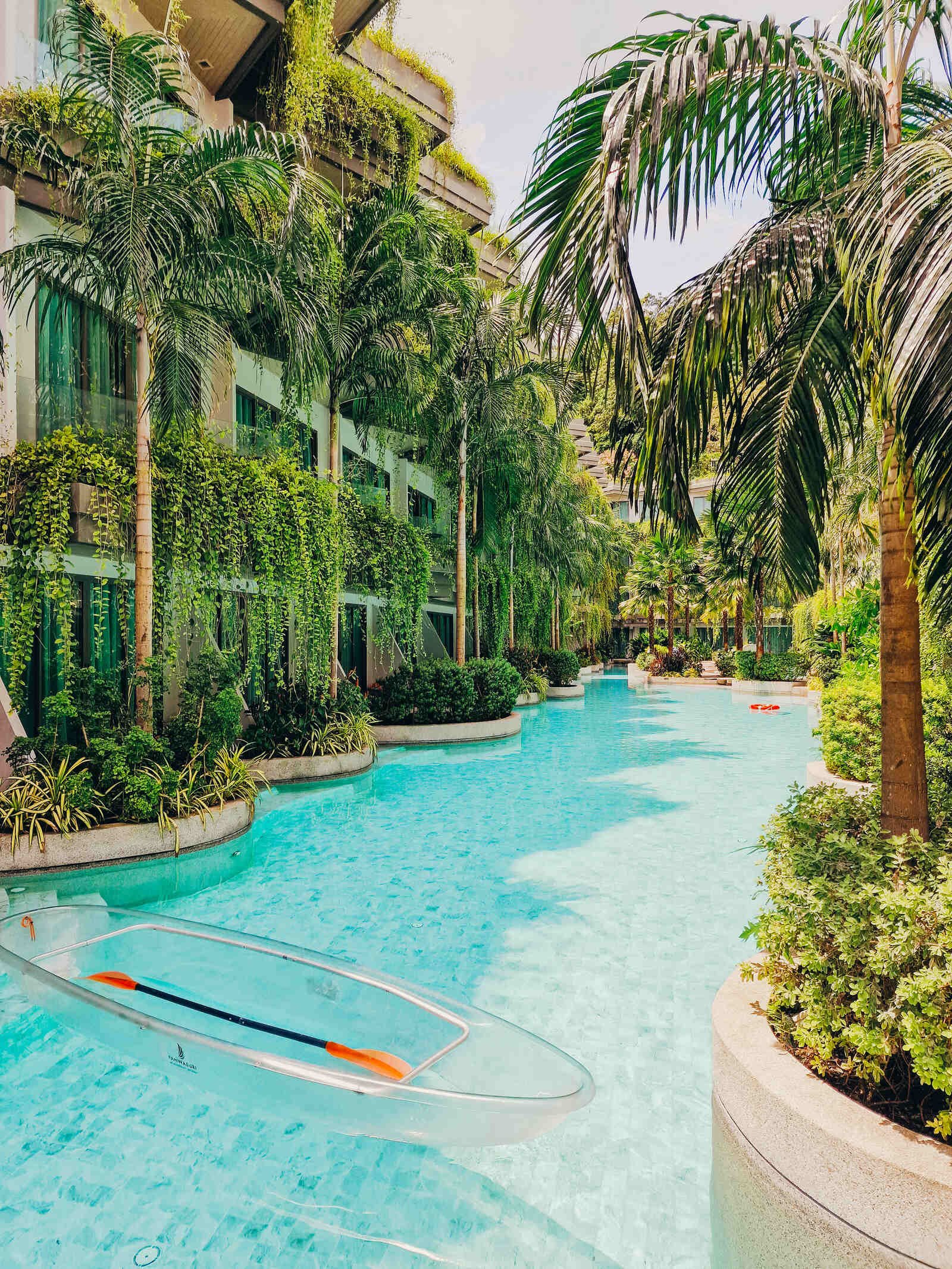 a hotel pool surrounded by greenery and palm trees with a glass bottom canoe floating empty in the pool