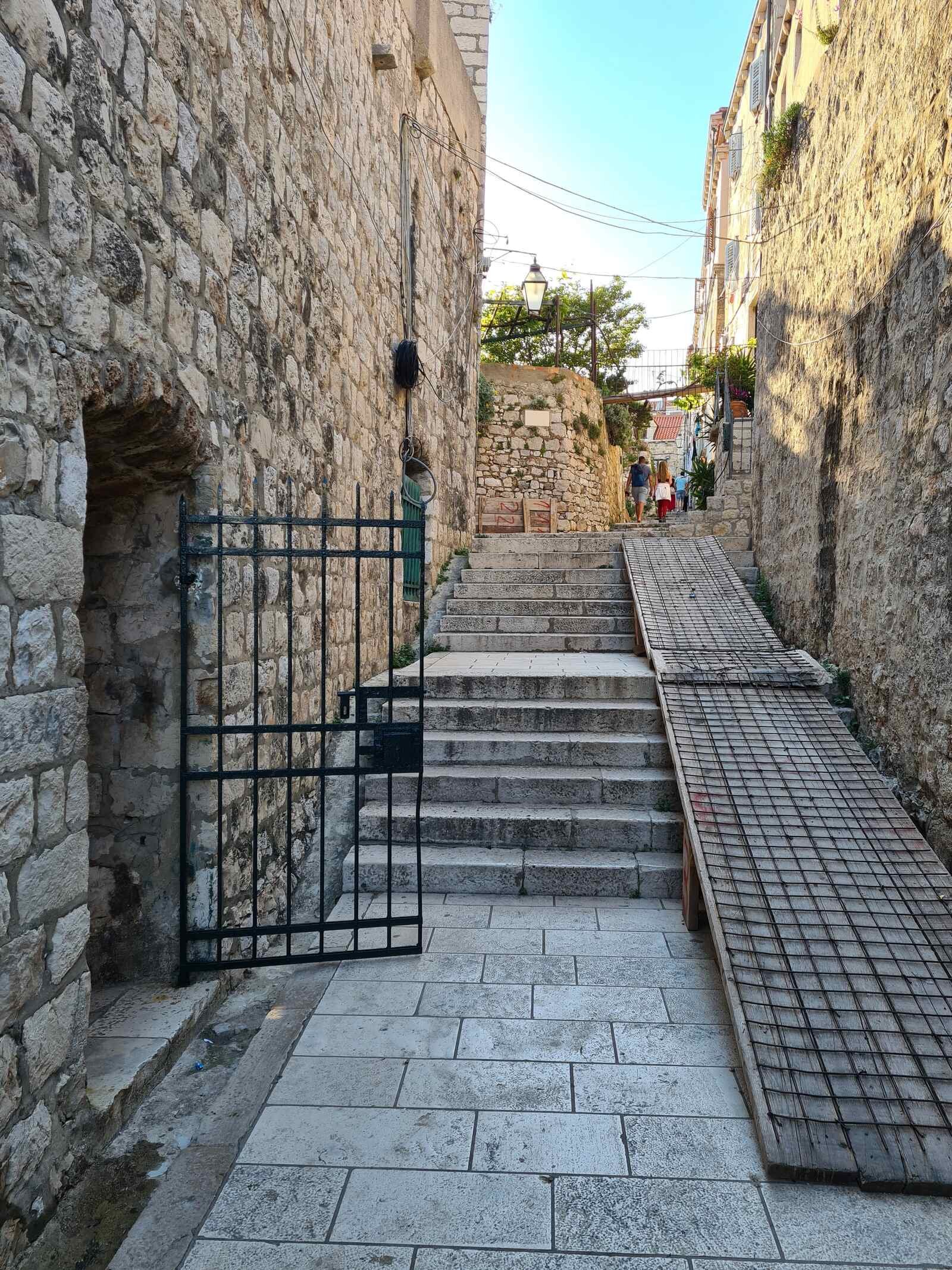 A cobble street with stone walls and steps leading up. A black iron gate is open in the wall on the left side