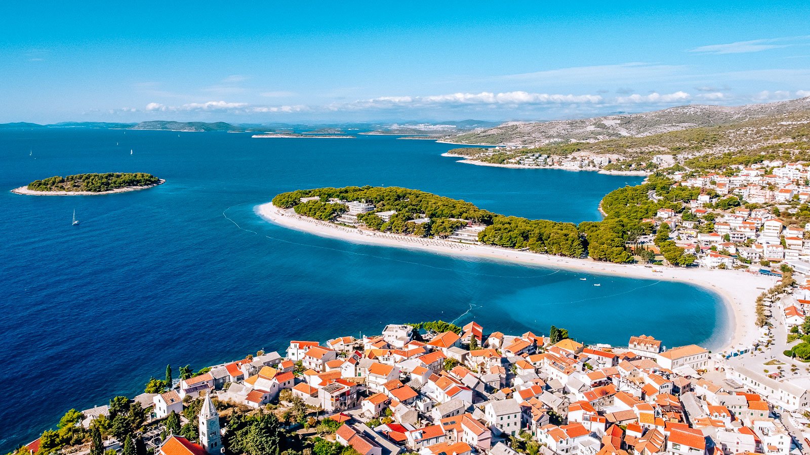 Drone shot looking along the adriatic coast in croatia, the sea below is bright blue with peninsulas of land sticking out into it covered in terracotta roof houses and green trees. Islands can be seen in the distance, it's sunny with blue sky