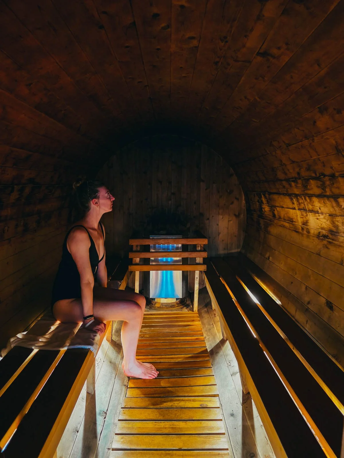 Helena in a black swinsuit sitting inside a long round wooden sauna