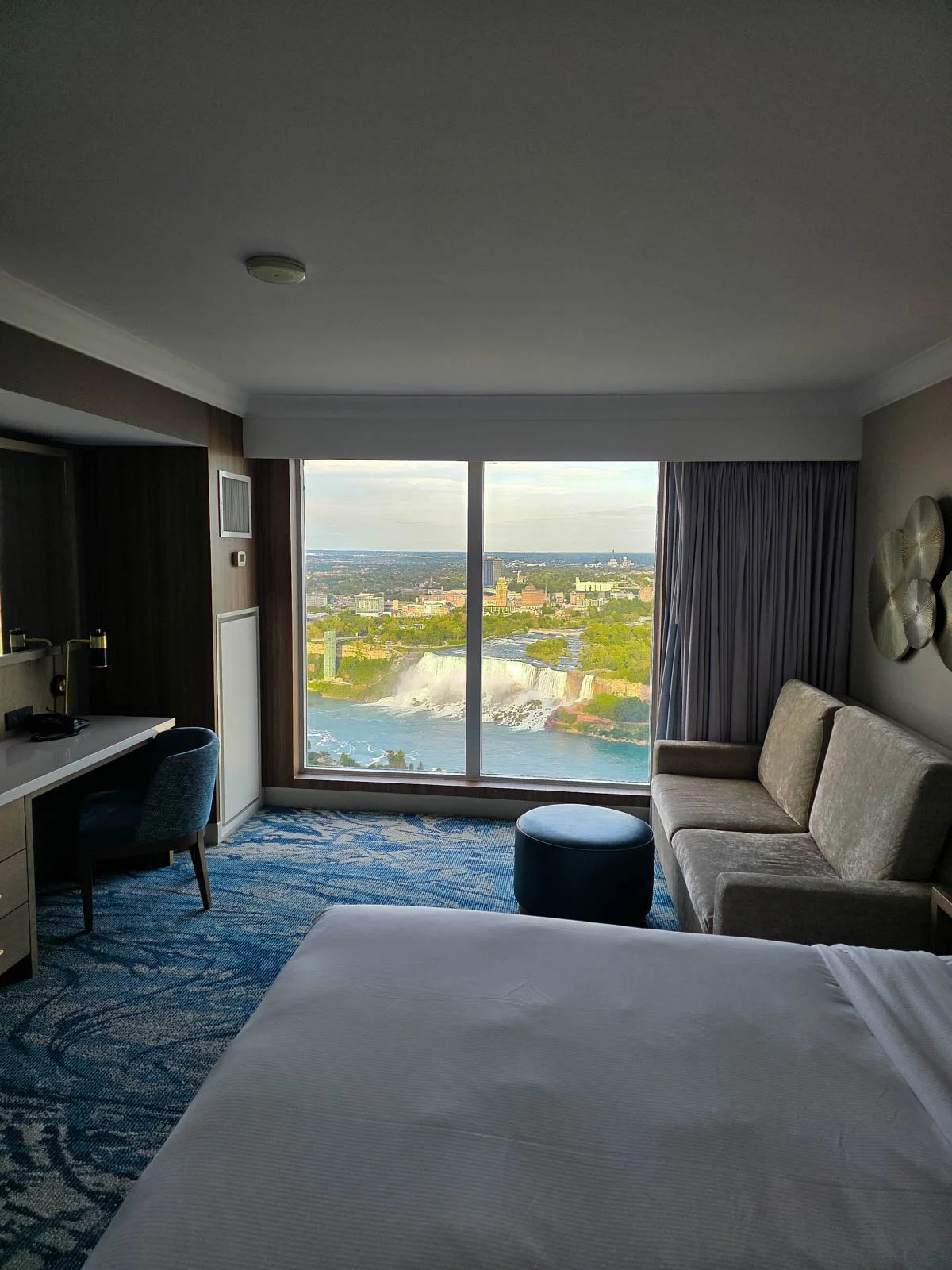 A view from inside a large hotel room looking across a bed and sofa to the floor-to-ceiling windows where a large waterfall can be seen on the other side of the river at Niagara Falls
