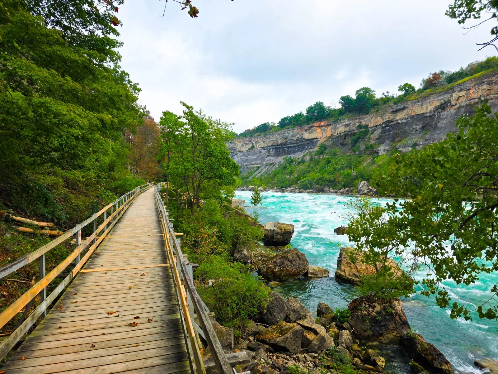 a wooden walkway along one side of a river gorge, trees are on the left, a turquoise fast flowing river on the right and the cliffside of the gogre is beyond, at the White River Walk in Niagara Falls Canada