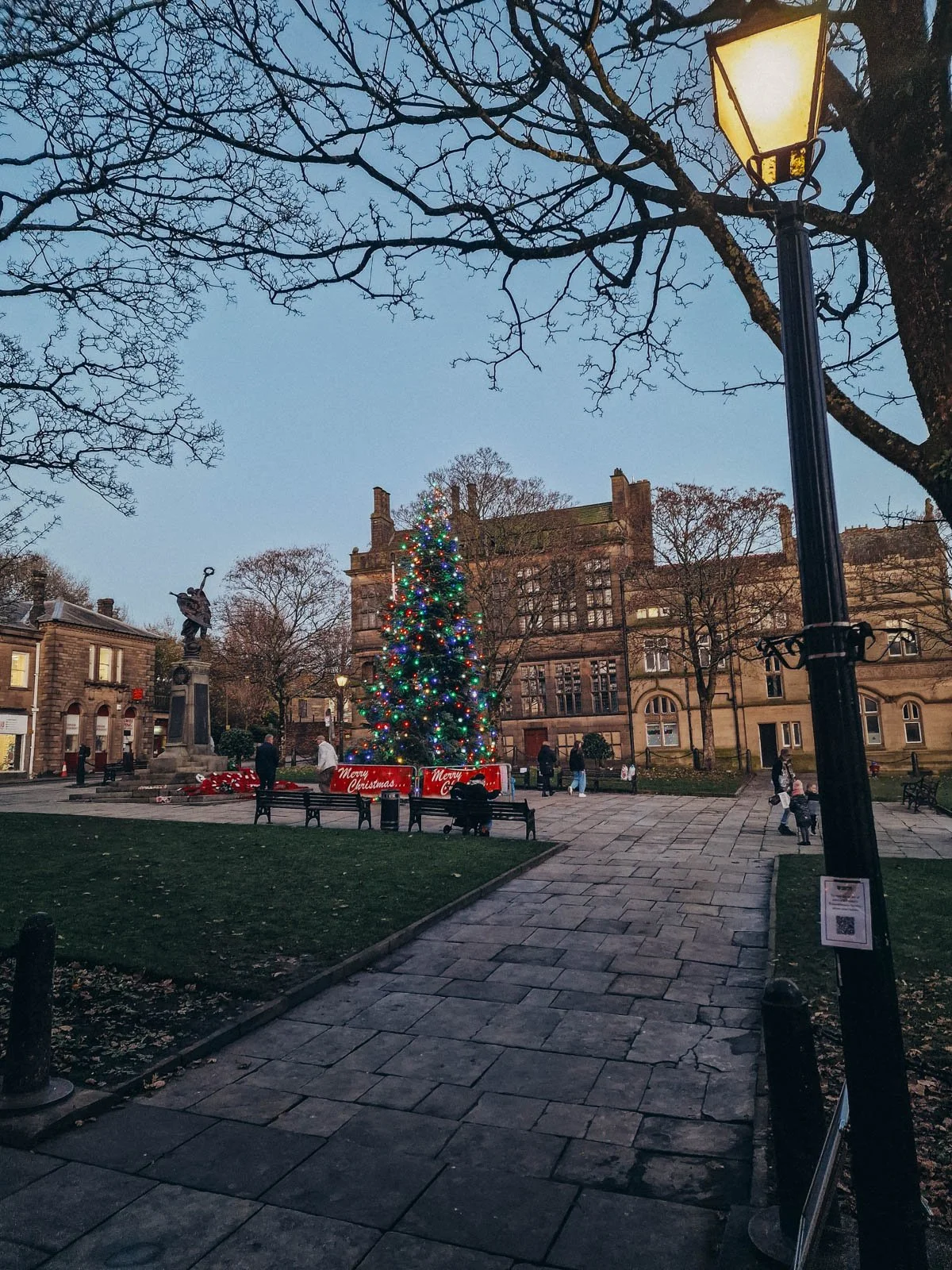 a town square in glossop reached by train from manchester, there are stone buildings on every side of the square which is pedestrianised with benches and people milling around and a christmas tree with multicoloured lights in the middle