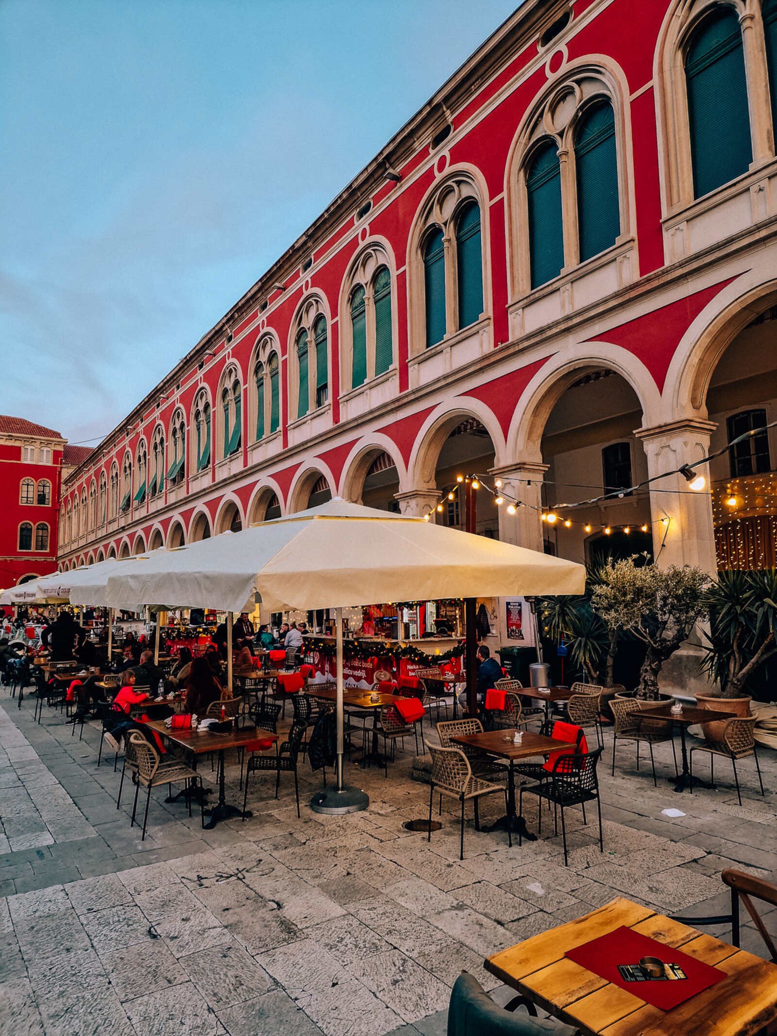 A large outdoor seating area of a restaurant with large parasols. A colourful old building with many  archways and windows