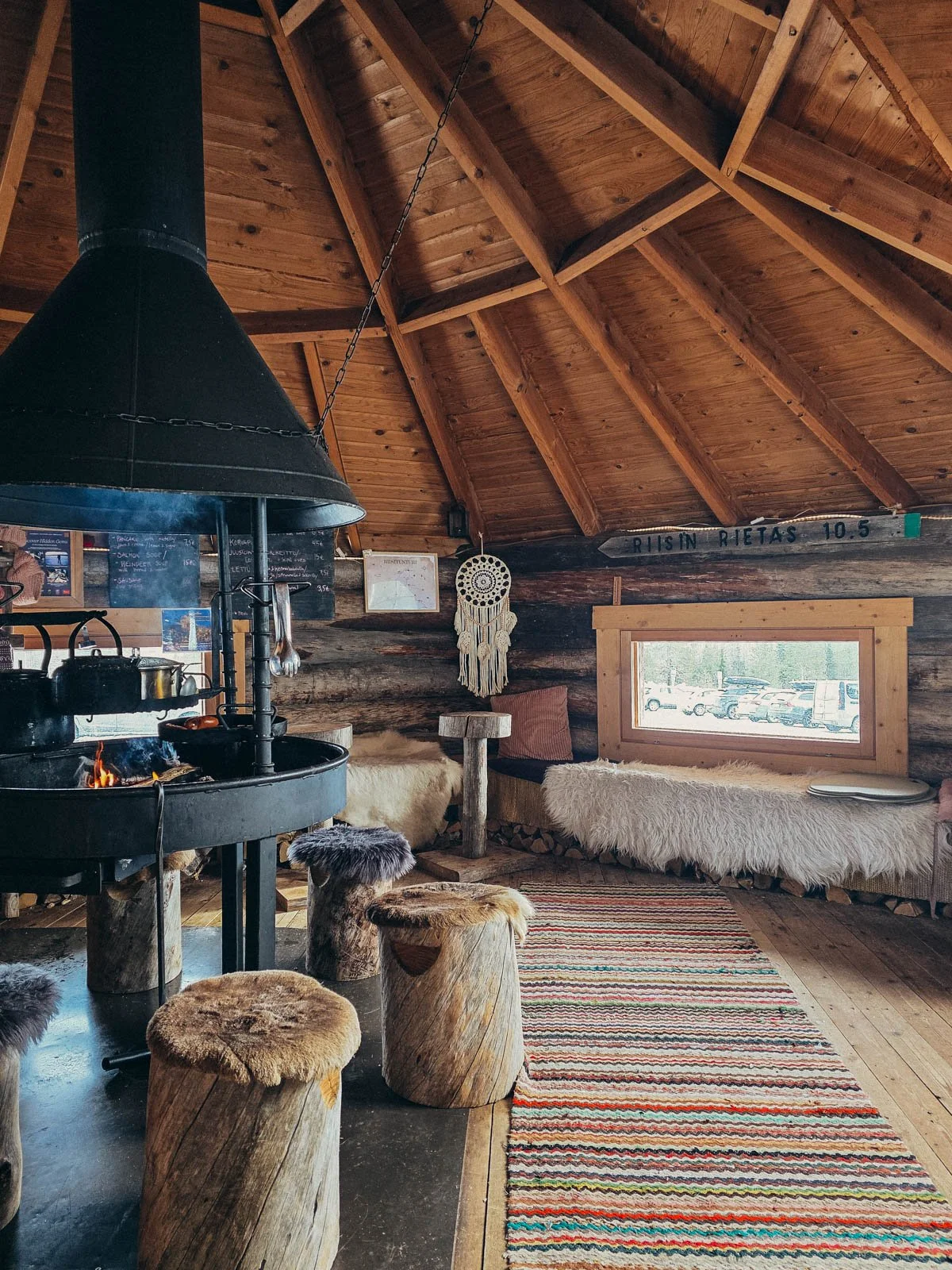 A small wooden hut cafe interior with a large black wood fire stove in the middle. Fur covered log chairs surround the wood stove