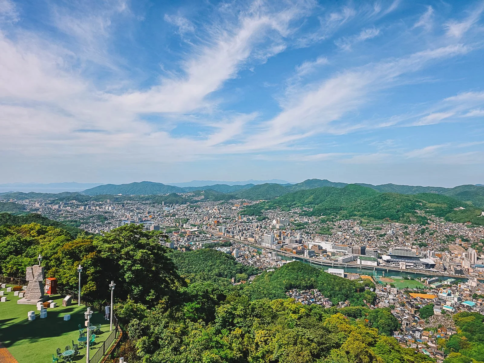 A panoramic view from a viewpoint looking down on the city of Nagasaki in a valley surrounded by lush green hills that stretch far into the distance, the sky is blue on a sunny day