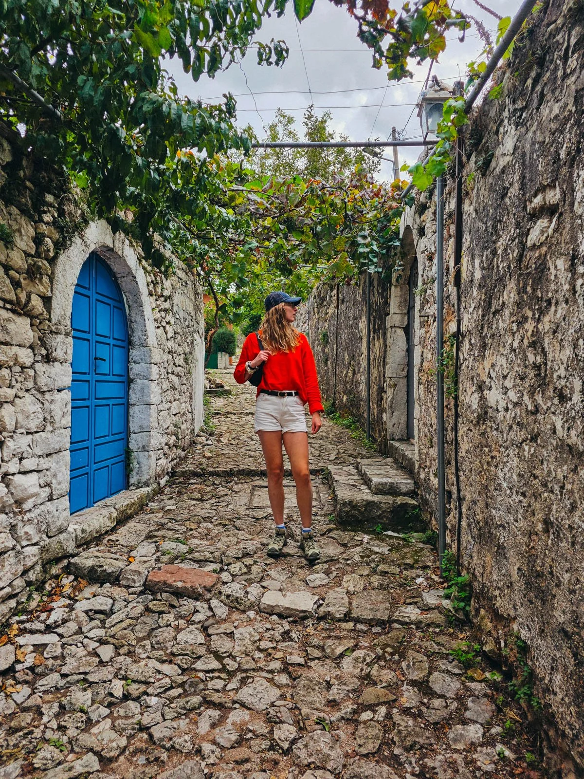 Helena in red standing in the middle of a stone path with stone walls and a blue door to her left