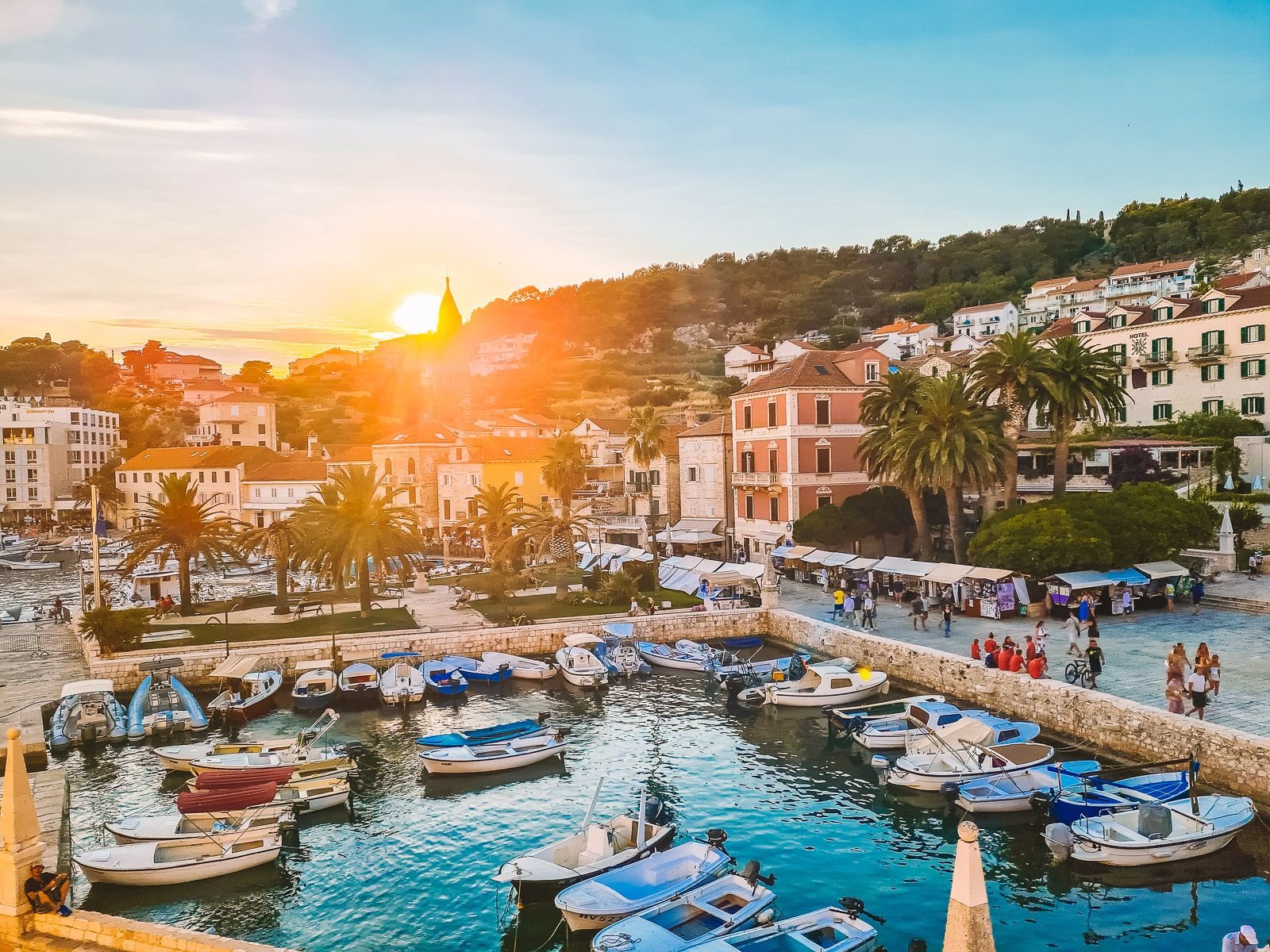 Looking down on a small harbour with boats in at sunset. Sun is setting between the hills in the distance and the hillside is covered in colourful houses with orange roofs on the island of Hvar, Croatia