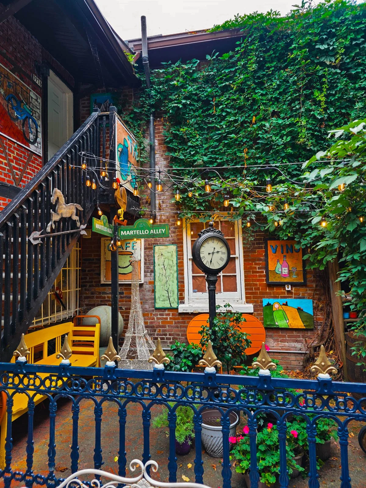 A colourful courtyard full of quirky items like a tree standing black clock, a street sign, a yellow bench, in the corner of a brick building half covered in ivy