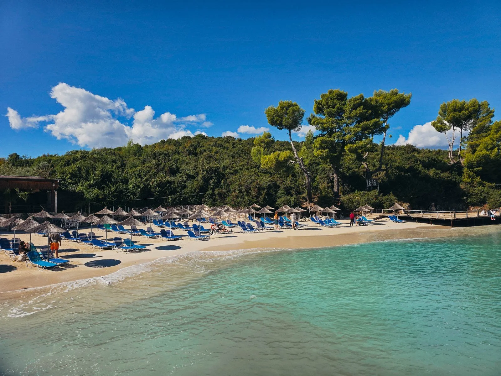 A  white sand beach with clear turquoise water. On the beach there are many beach chairs and umbrellas with trees beyond the beach