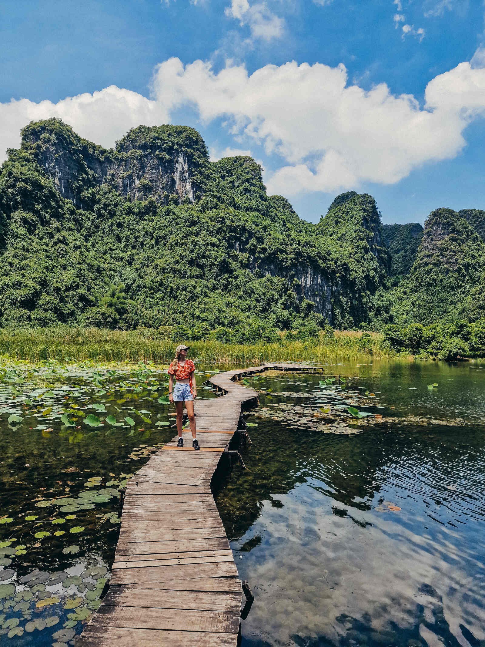 Helena standing on a wooden walkway on the water with green lilypads and lush cliffs in the background