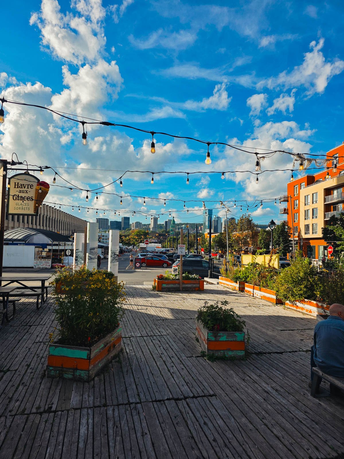 A wooden deck with plants and string lights, beyond it is a market hall and the city skyline on Montreal beyond