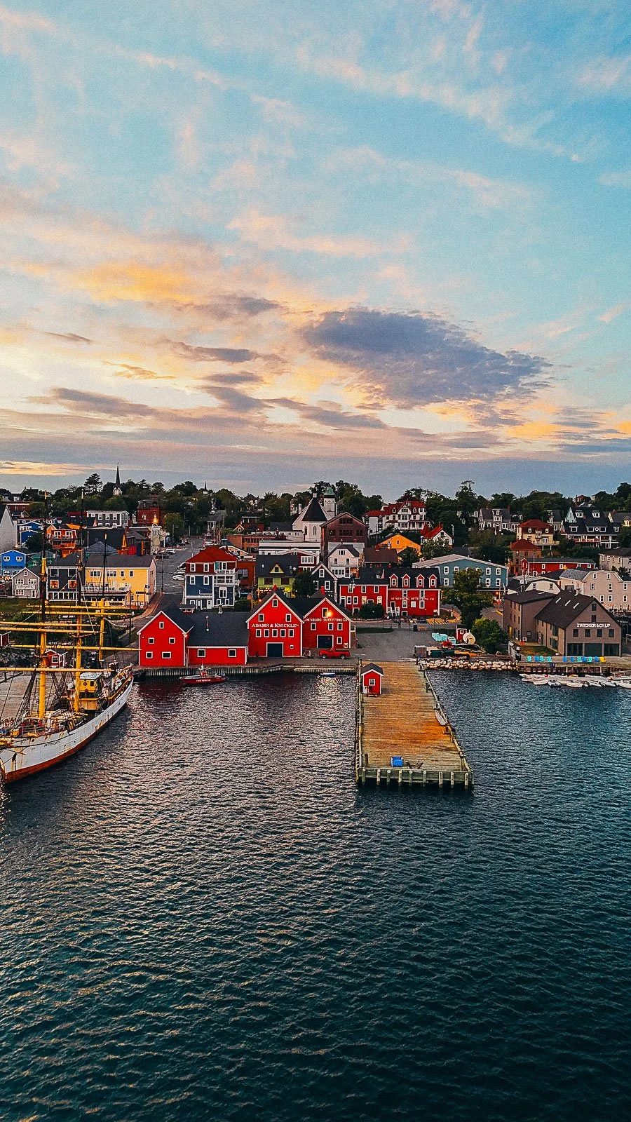a drone shot from above the water, looking down at the colourful red and yellow waterfront buildings in Lunenburg with a pier stretching out into the water at sunset
