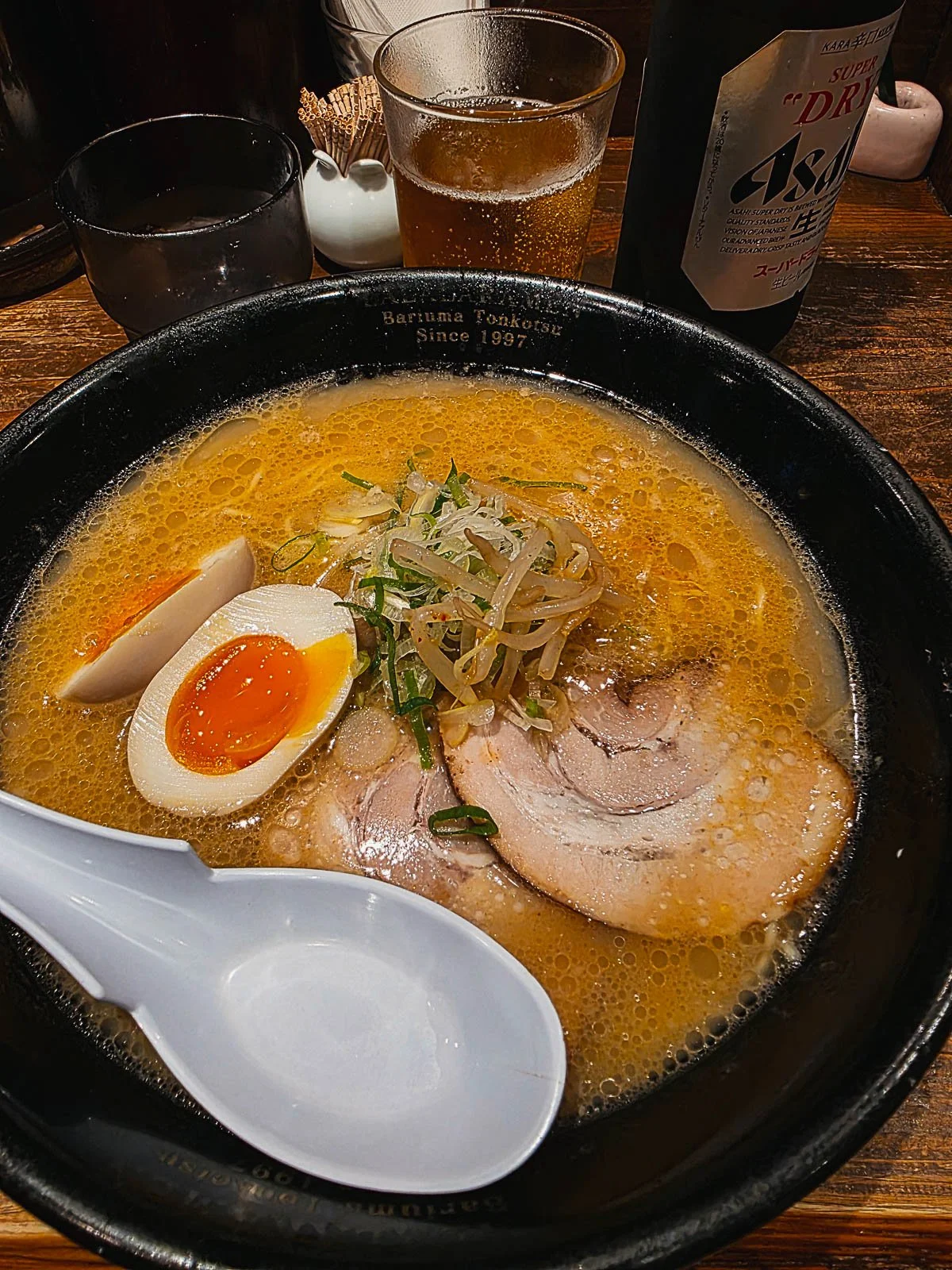 a black bowl full of ramen with meat, bean sprouts, soft boiled egg and a white spoon