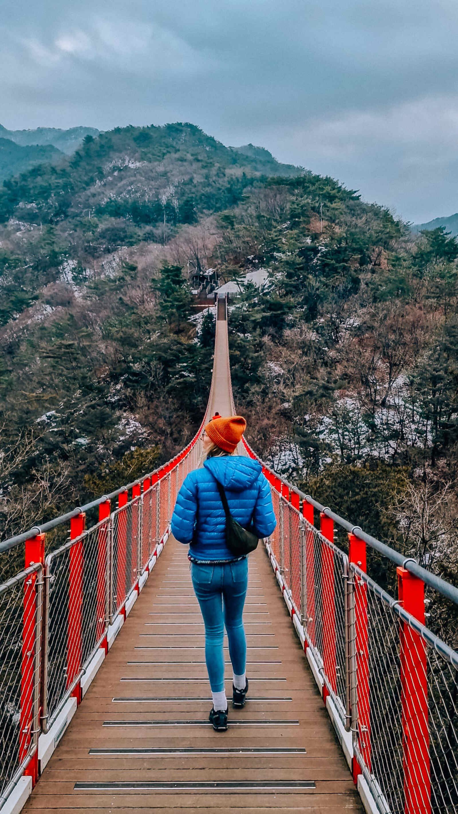 a woman in a blue coat walking across a red suspension bridge towards a hill with some snow on it on the other side.