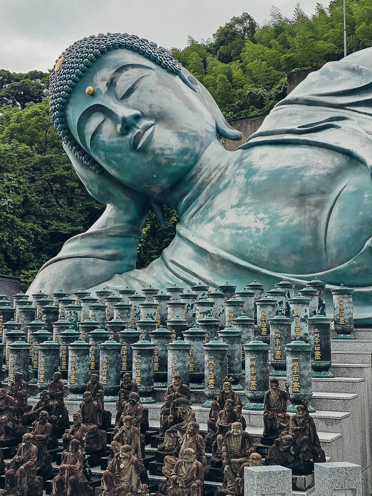 A large blue-green reclining buddha at Nanzoin Temple in Kyushu Japan with many small Buddha statues in front