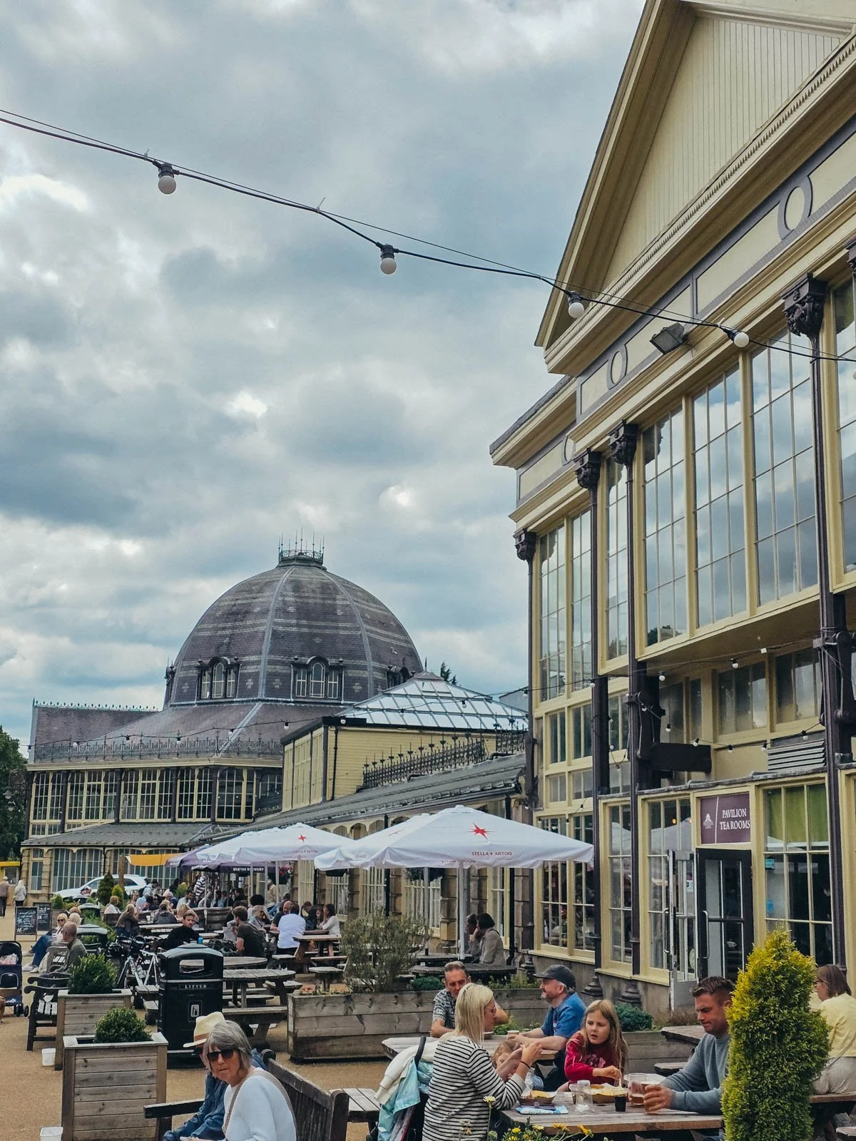 Many people sitting outside on benches underneath parasols outside an old fashioned glass pavillion