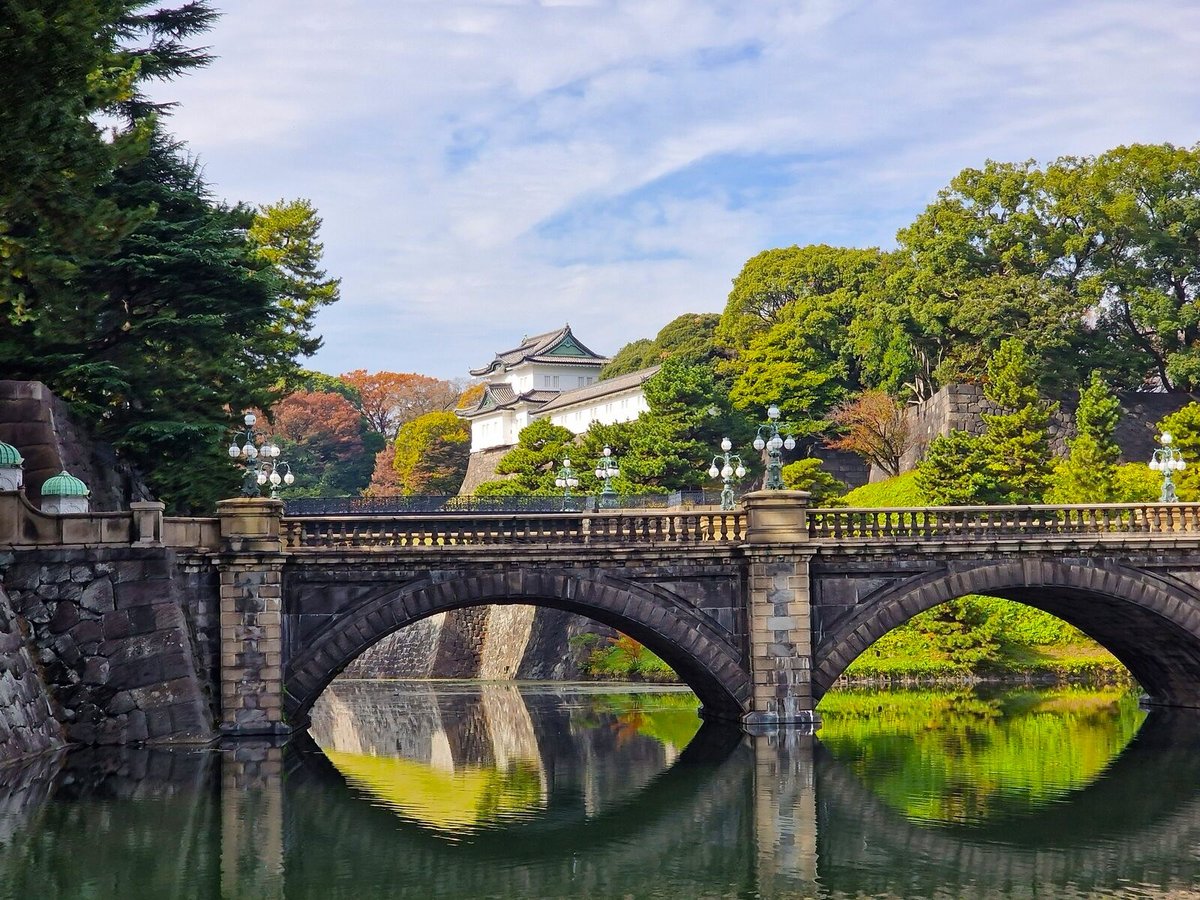 a stone bridge with the roof of the imperial palace in the distance obscured by some green trees