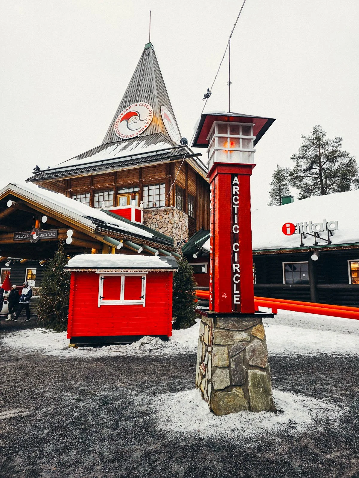 A large red post marking the arctic circle with a large pointed wooden building in the background surrounded by snow and gravel at Santa Claus Village in Rovaniemi