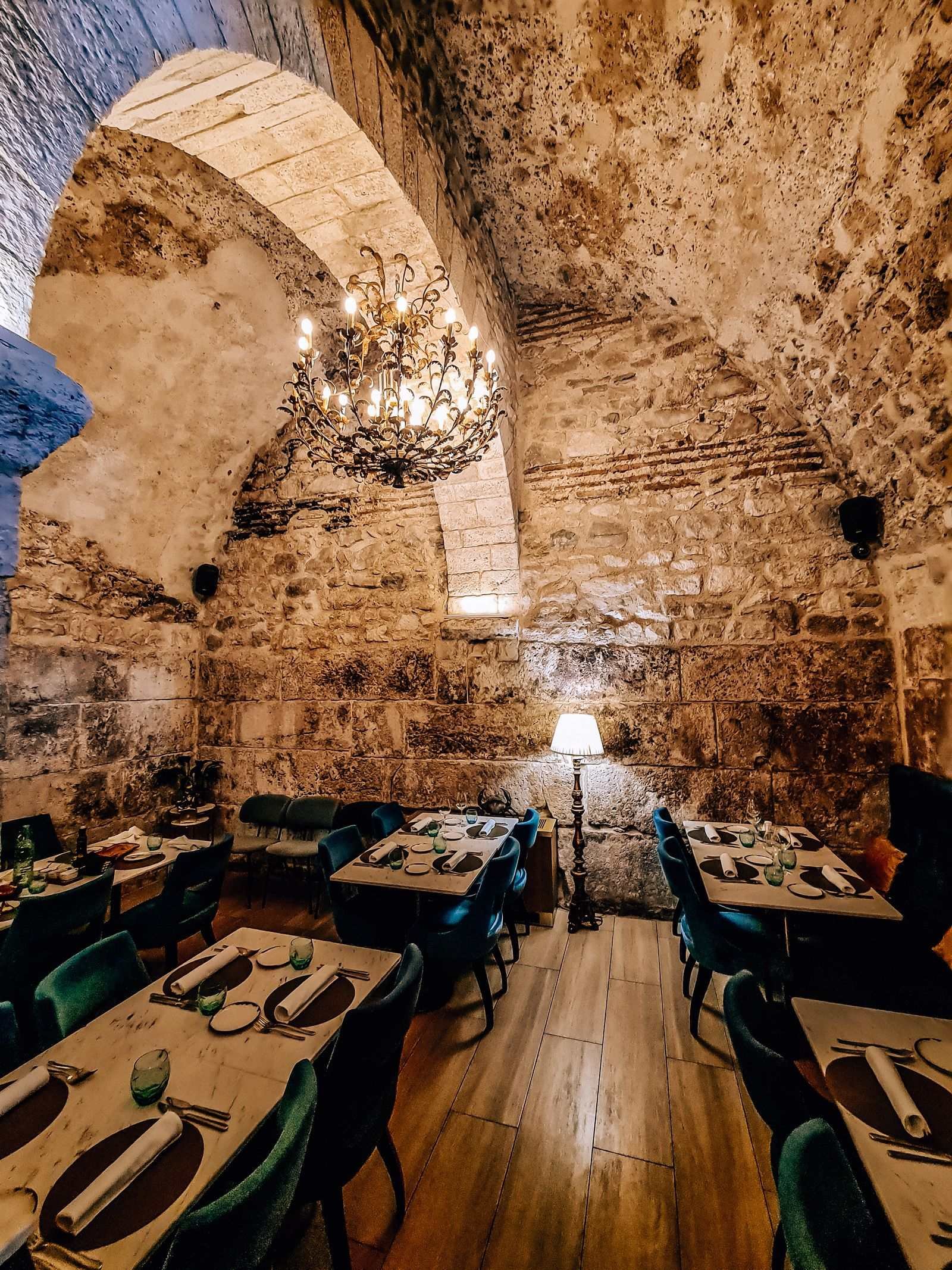 The the old arched stone walls and ceiling of a restaurant with many tables and a chandeliarlier hanging in the centre