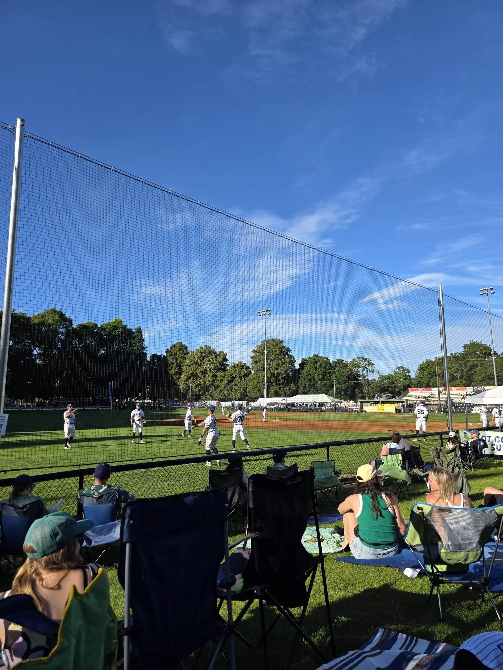 People sitting on game chairs behind netting with a baseball game going on, on the field with many players dressed in white