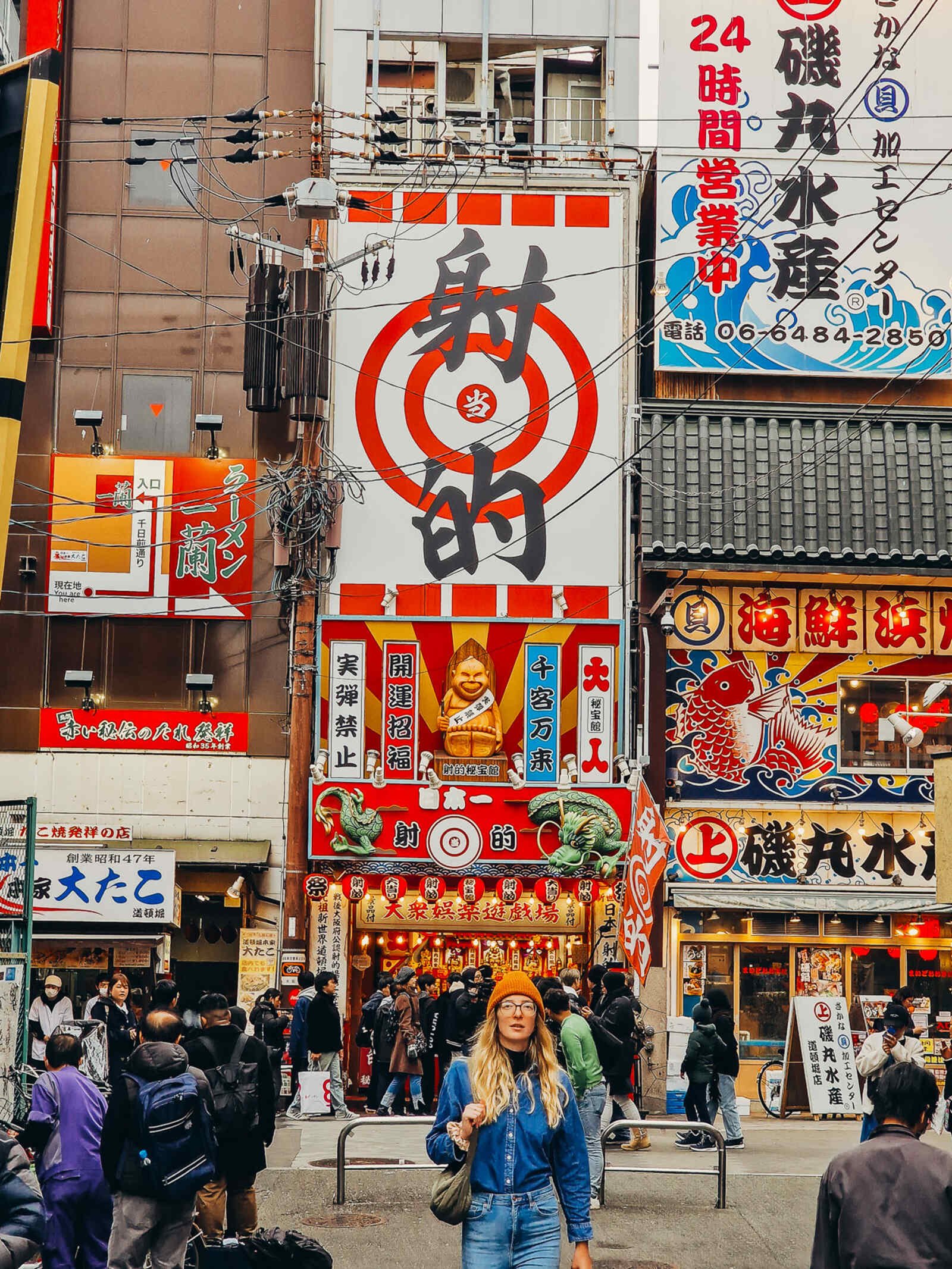 Helena in a blue denim shirt and jeans walking down a colourful street covered in bright signage, shop fronts, lanterns, in yellow and red with black Japanese writing in Osaka's Dotonbori district
