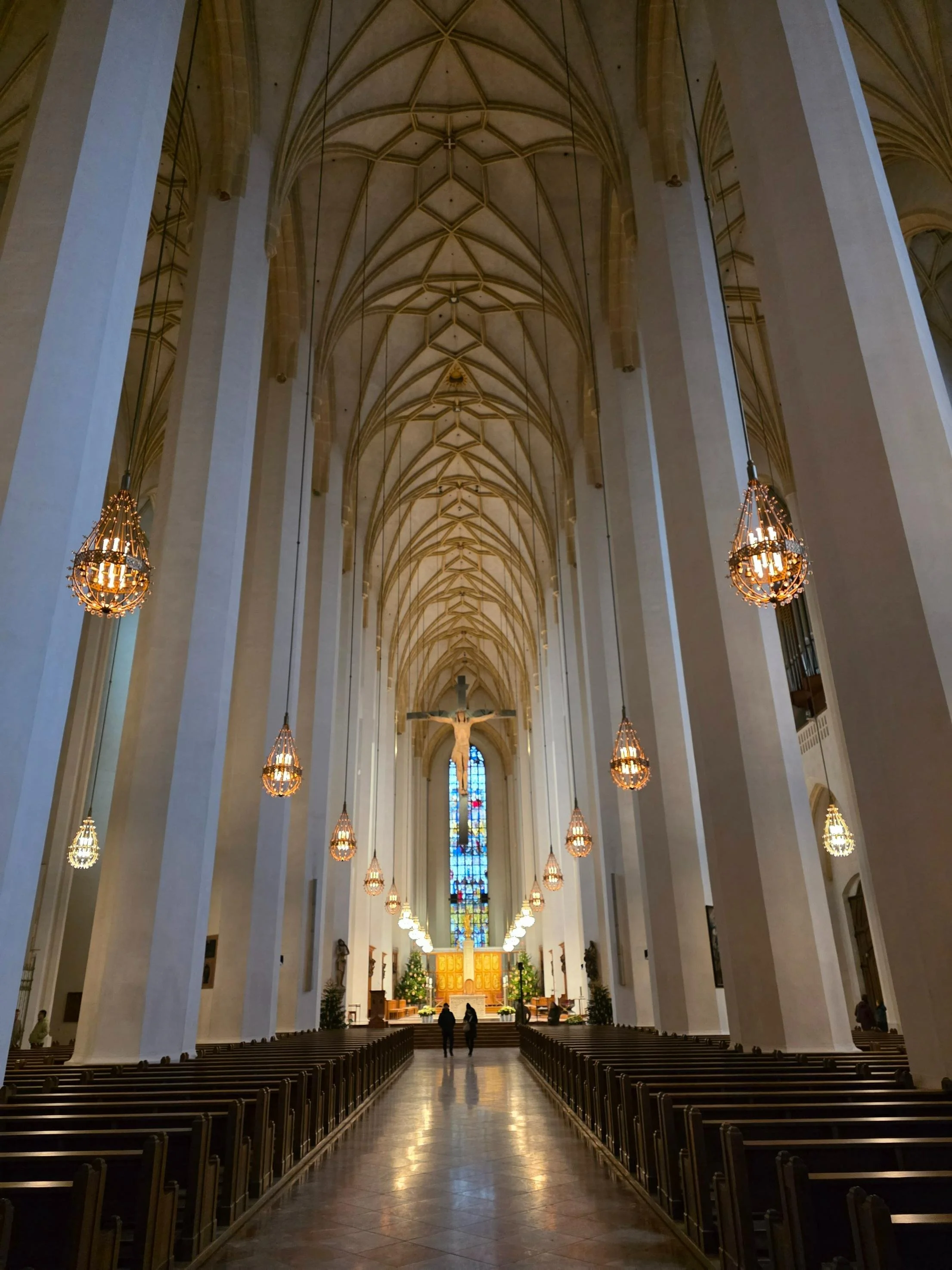 The large high ceiling interior of a church with many tall thick white columns, a large cross, dark wooden pews, and stained glass windows at the far end of the church