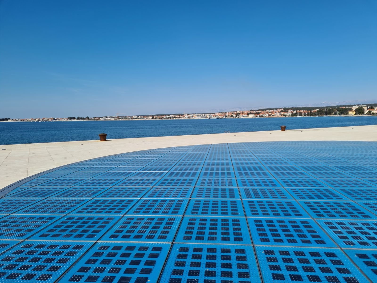 A large blue circular solar panel that is walkable near in the middle of a stone boardwalk along the water