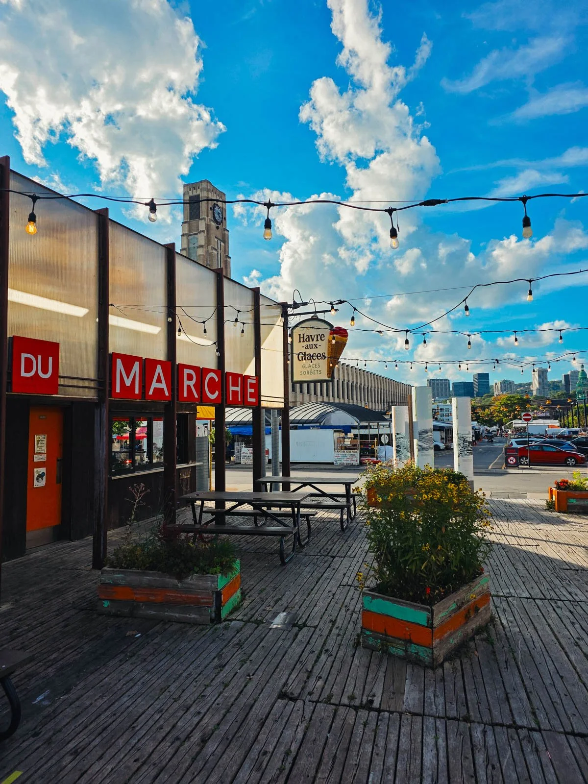 A wooden deck with plants and string lights, and a building with a sign that reads "marche", beyond it is a market hall and the city skyline on Montreal beyond