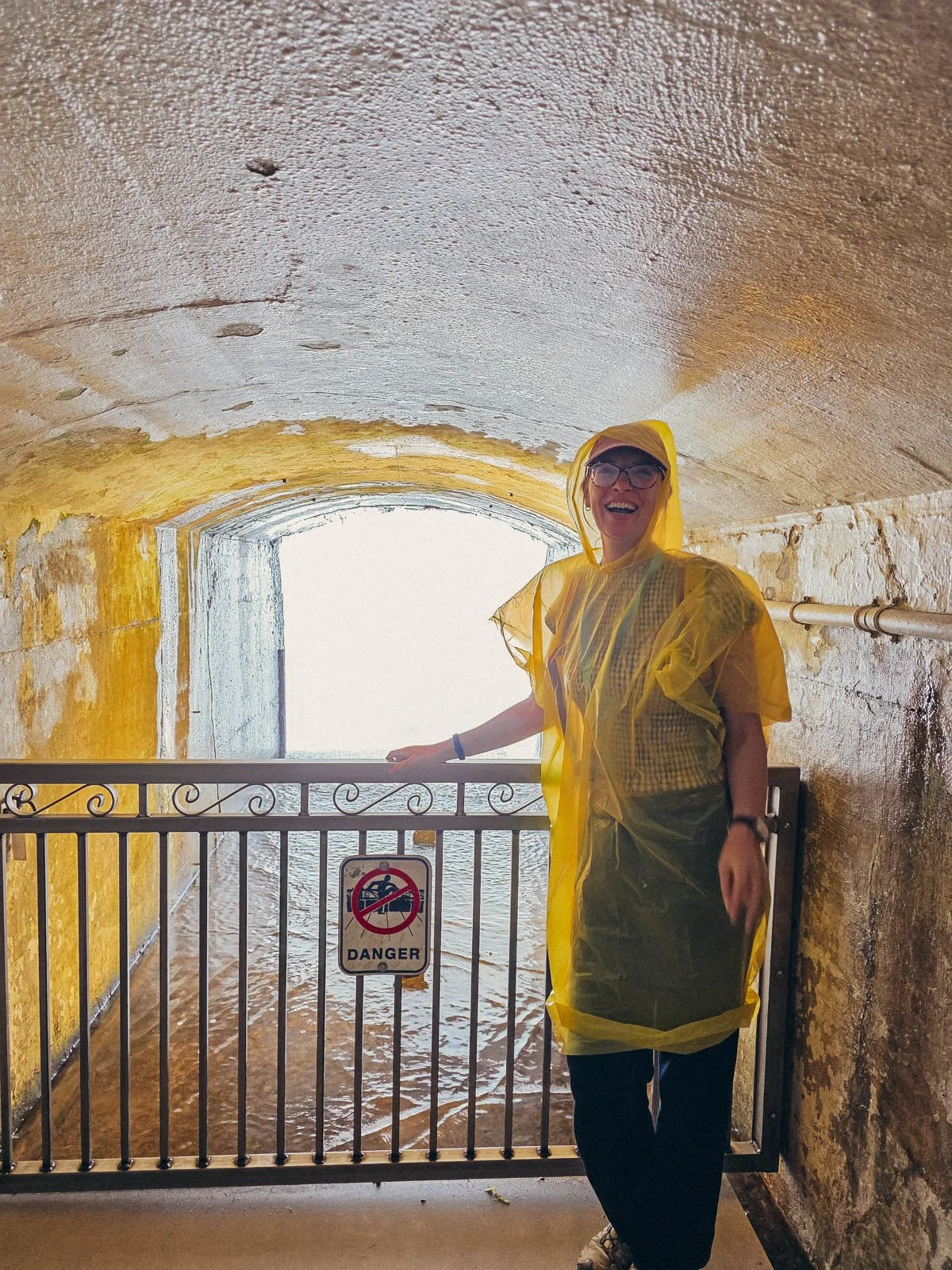Helena in a yellow poncho with the hood up standing in a tunnel with water on the floor and she's laughing at the camera, at the Journey Behind the Falls attraction at Niagara Falls
