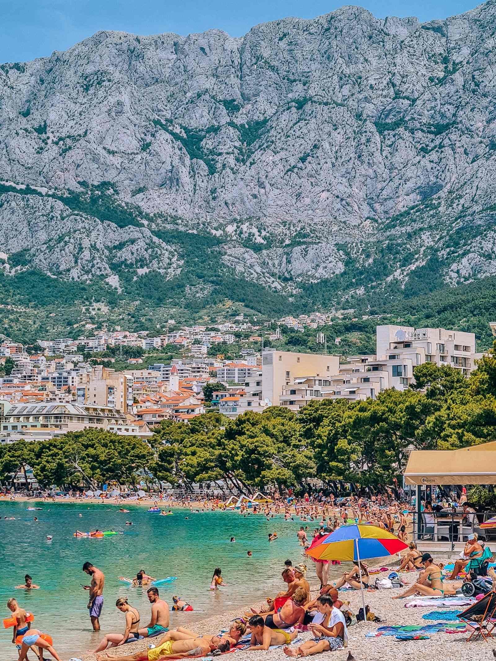 A packed beach with emerald green water, lined with trees and a town in the distance on the mountainside. A huge mountain dominates the background of the photo
