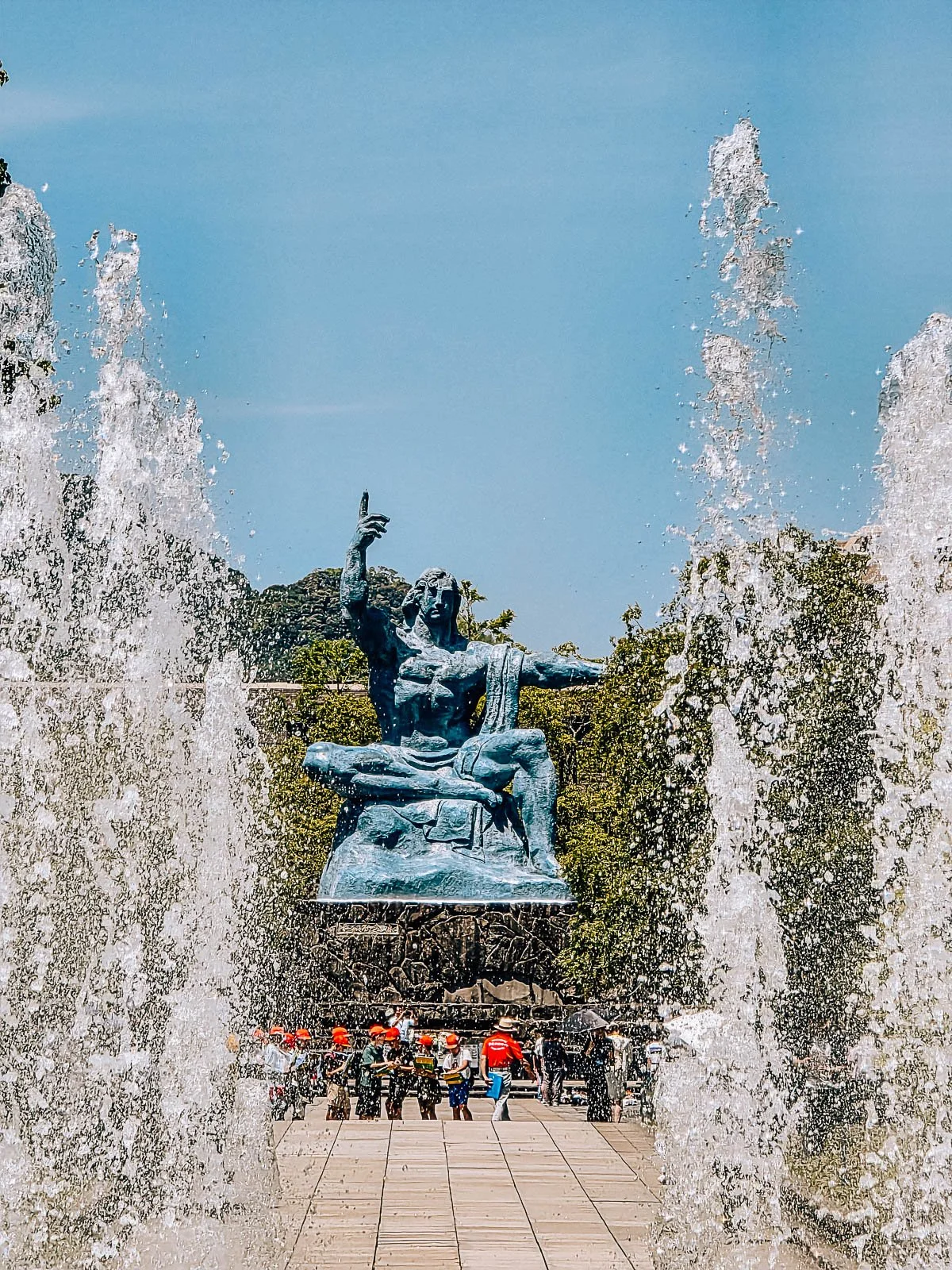 A large blue statue of a muscular man drapped in cloth with one hanf in the air and the other out to the side, a fountain spraying up in the foreground