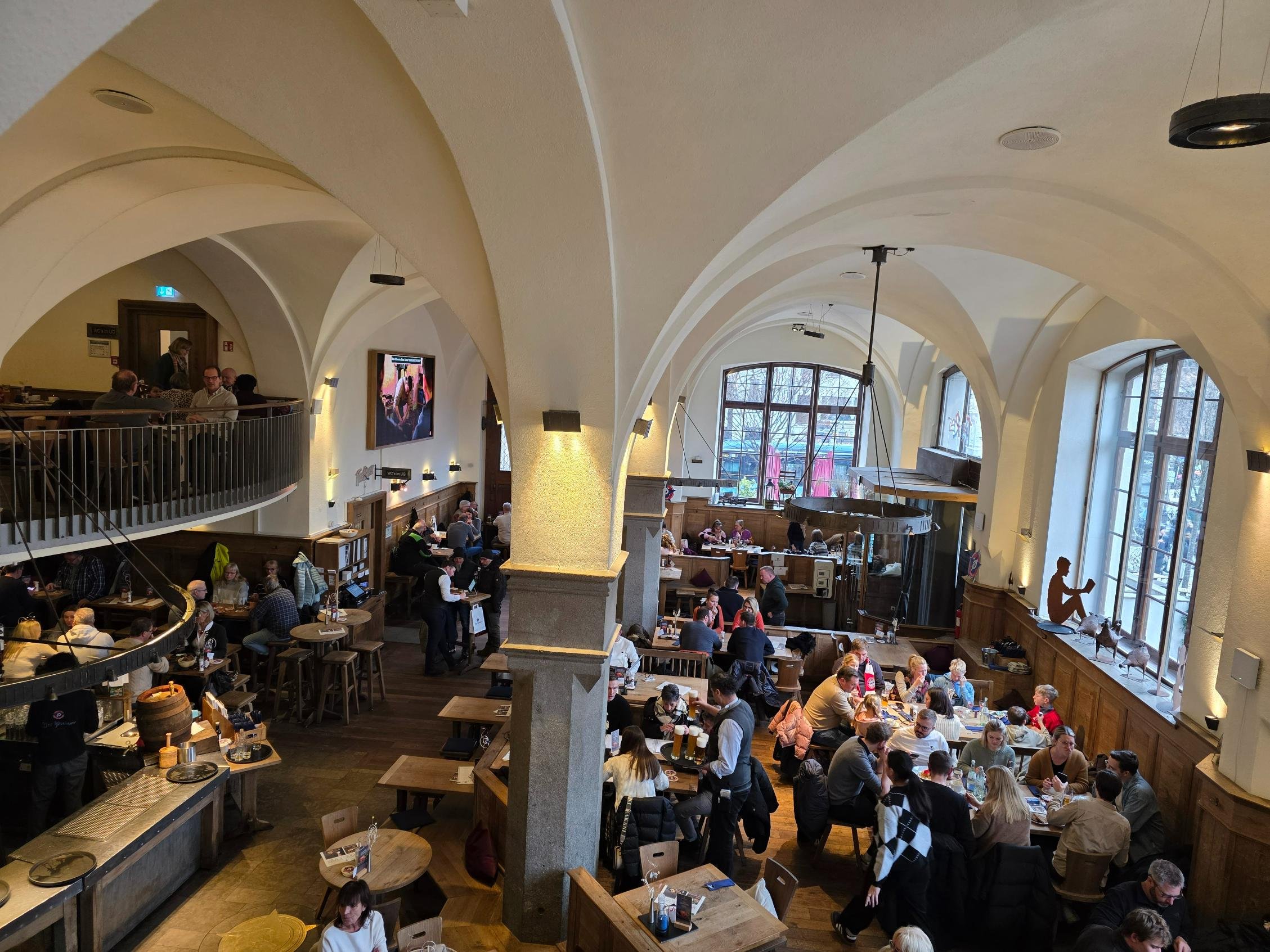 The view  from a balcony looking down ata large beer hall with many tables of people eating and drinking