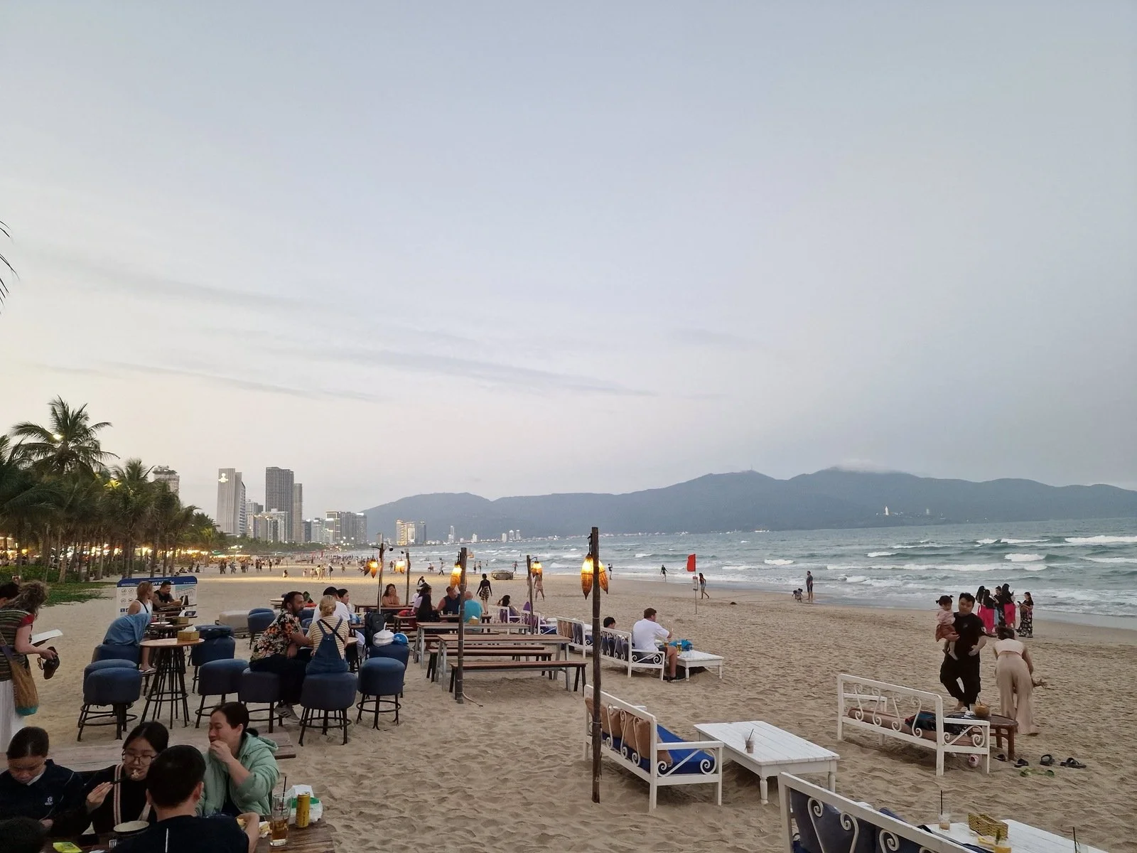 Many chairs and tables outside on the beach at a beach bar with the sea stretching out into the distance at sunset and mountains in the distance