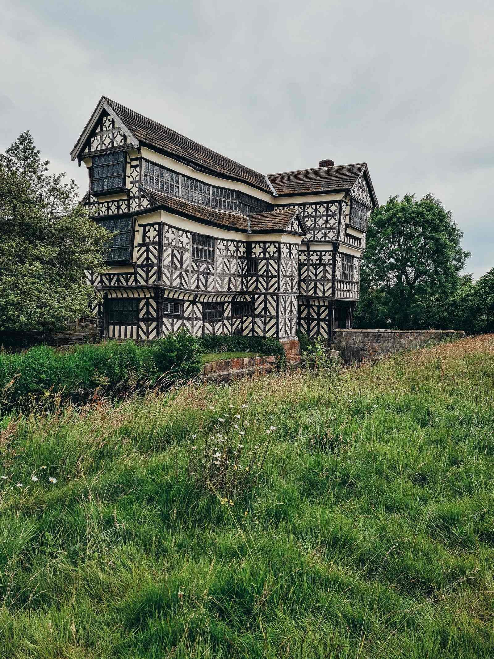 a large 3 storey black and white timber framed tudor mansion that leans outwards, situated in a grassy meadow