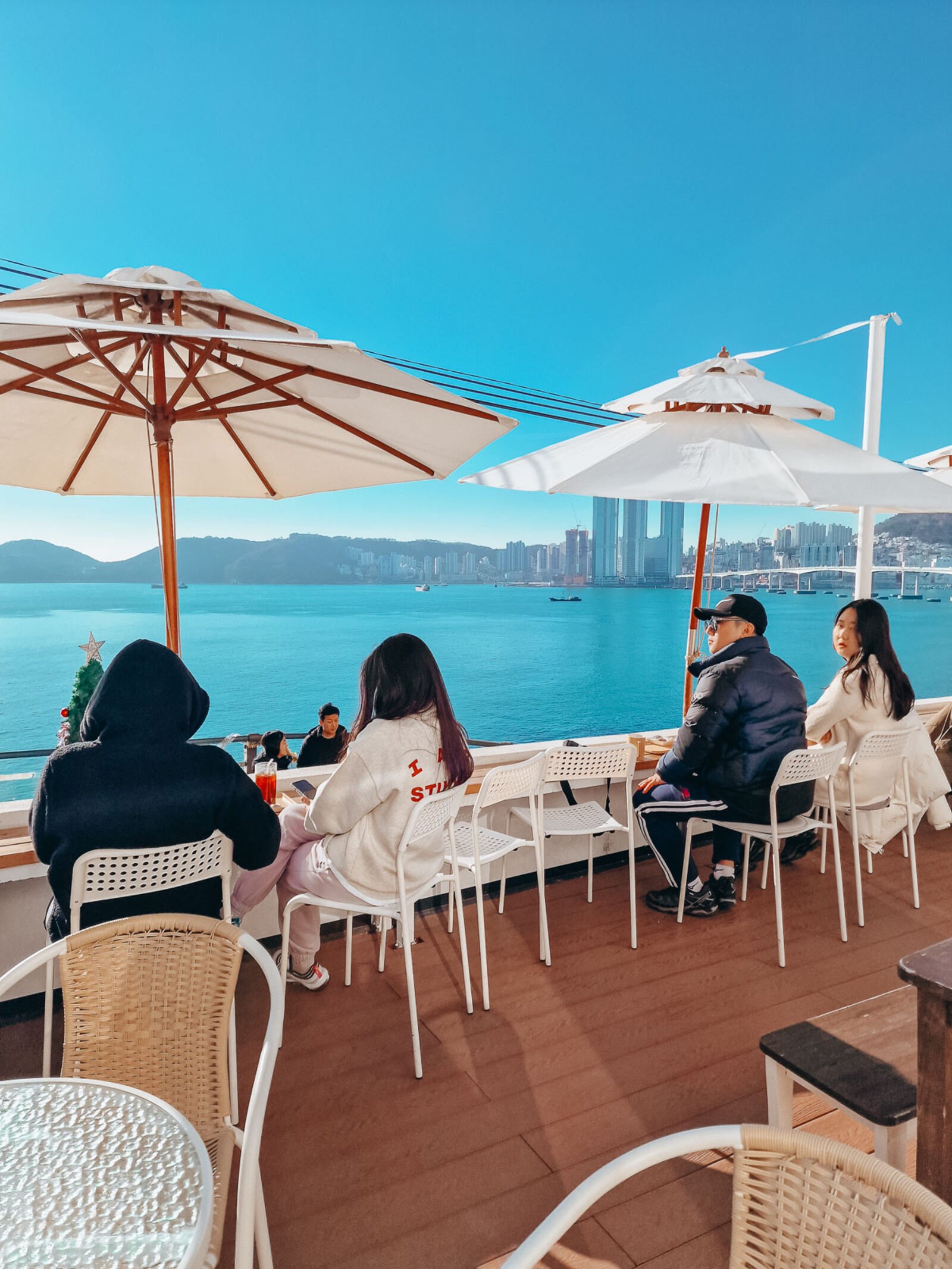 four people sit underneath parasols on a roof deck looking out across a bay of water on a sunny day with clear blue skies