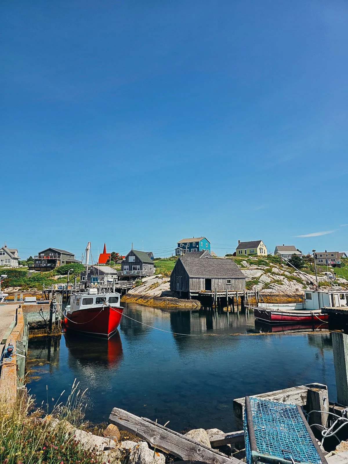 The cove of Peggy's Cove on a sunny, calm day. A red hulled boat is moored with many saltbox houses dotted on the hill on the opposite side of the harbour