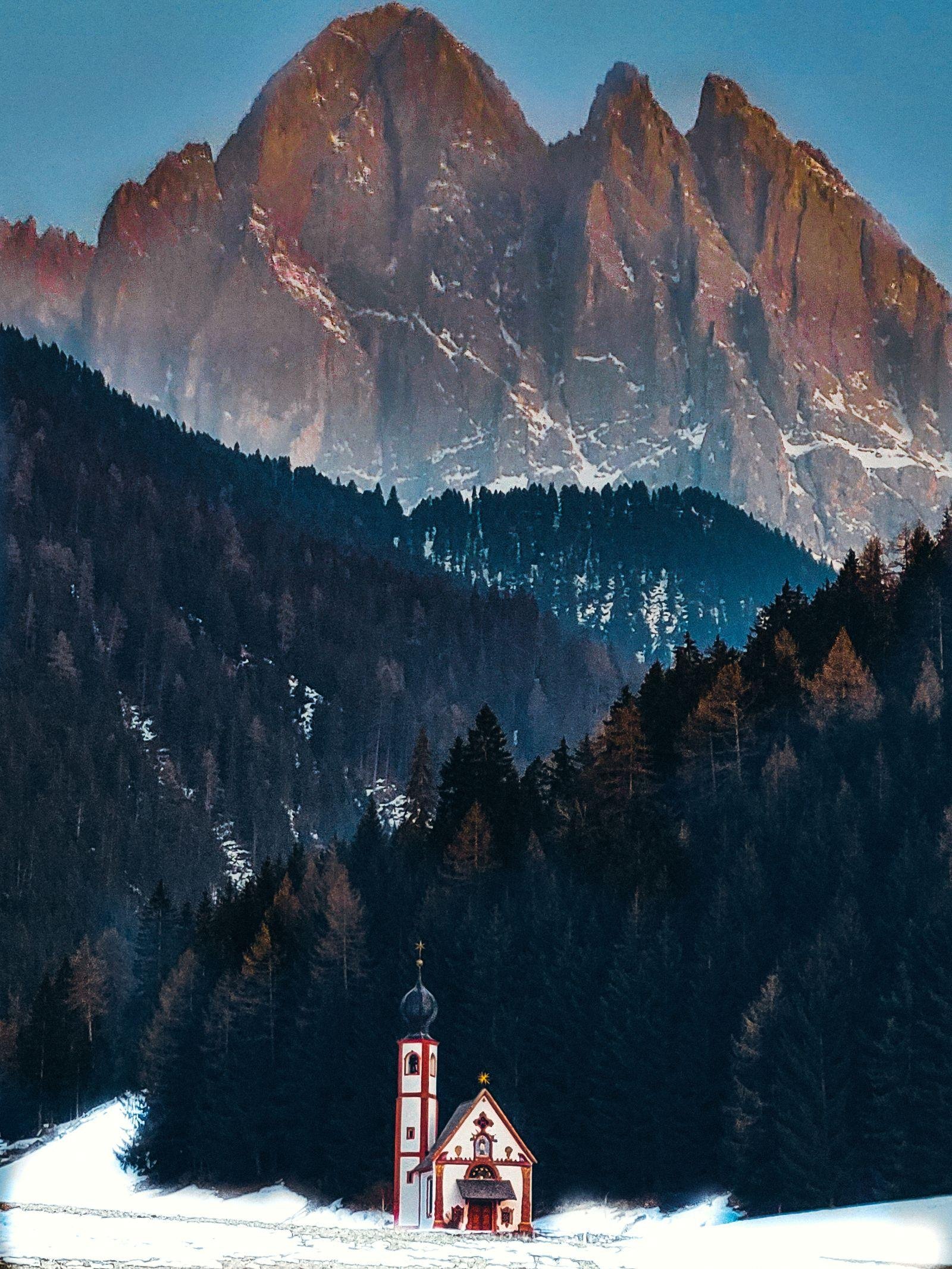 picturesque red and white church in the snow with dark trees behind and jagged mountains in the background glowing red and sunset in val di funes, Italy