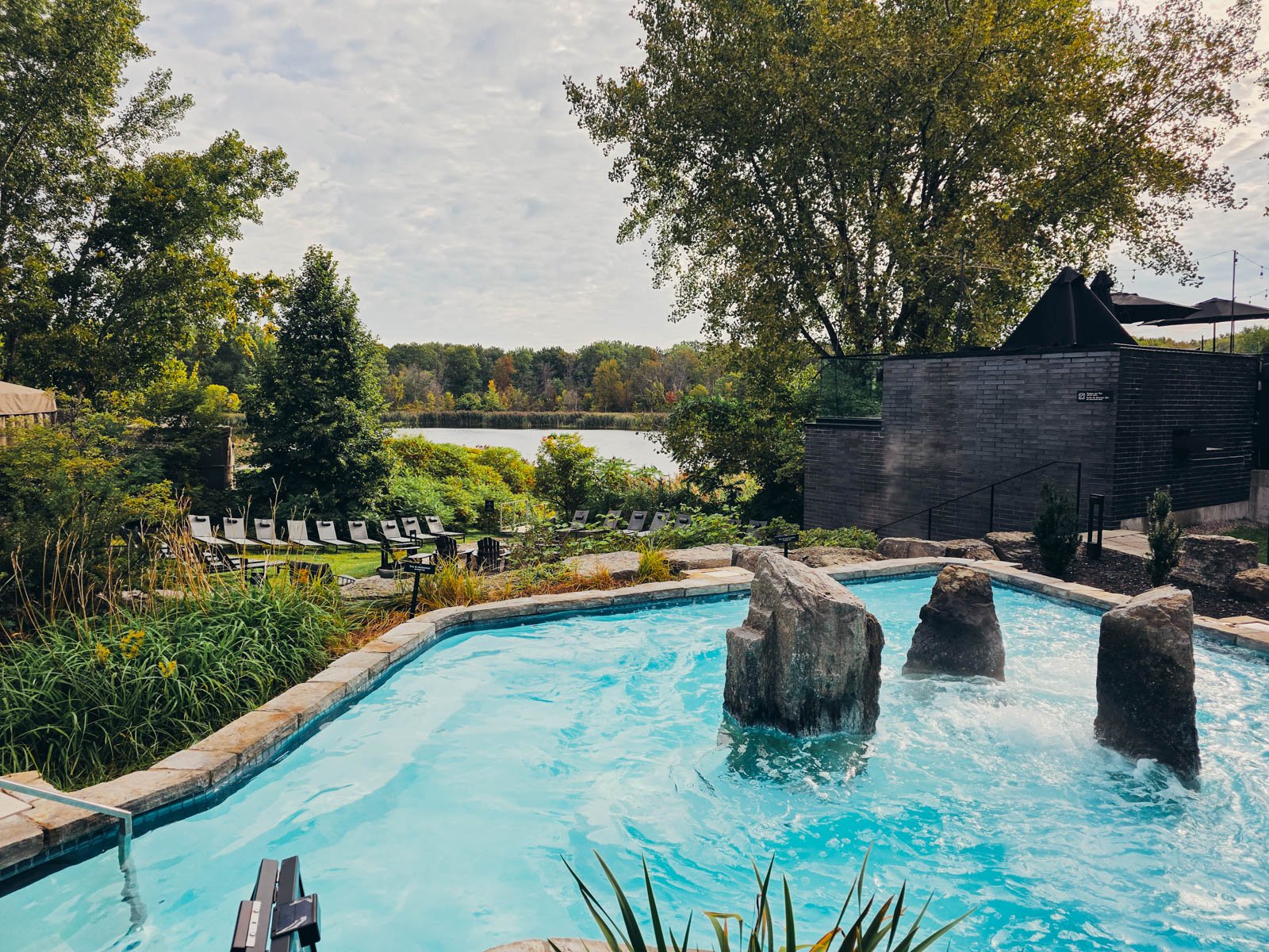 An outdoor spa with a bubbling blue spa pool with three large rocks in it. Beyond is a lawn with sun loungers and a lake is visible in the distance