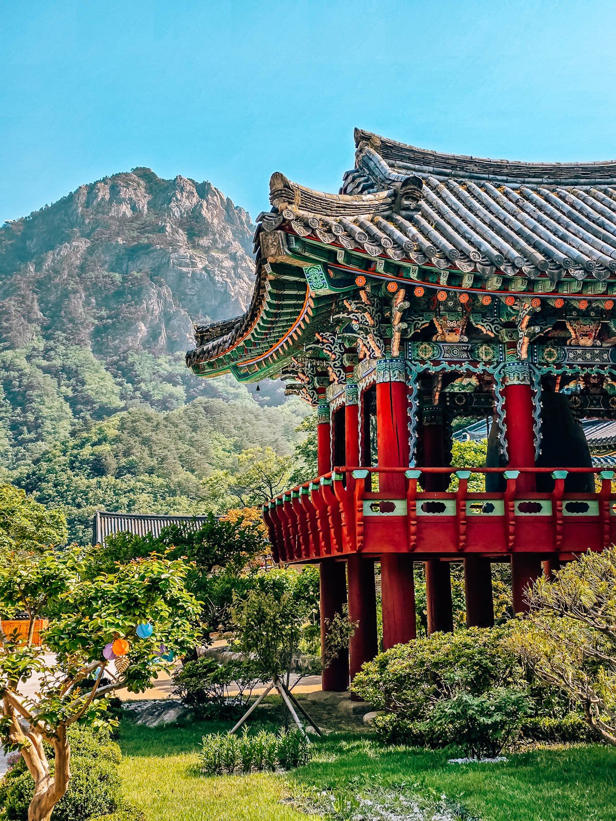 An elevated temple with red pillars and stilts and an ornate roof with green, blue and red colours. Beyond is the rocky mountain range of the national park