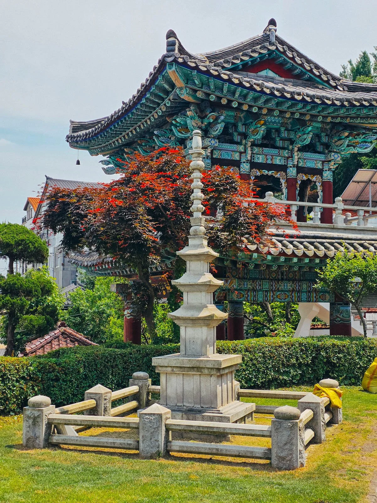 A large colourful turquoise temple building with a stone structure in the middle of a lush green lawn in front of the temple building