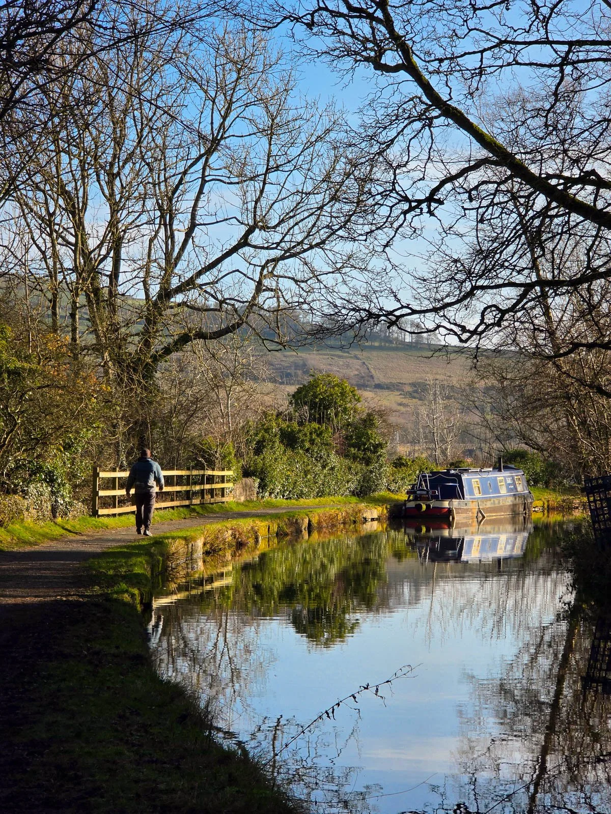A person walking away along a canal path with the canal curving away around the corner, one narrowboat is moored along the bank of the canal with warm sunight and blue skies