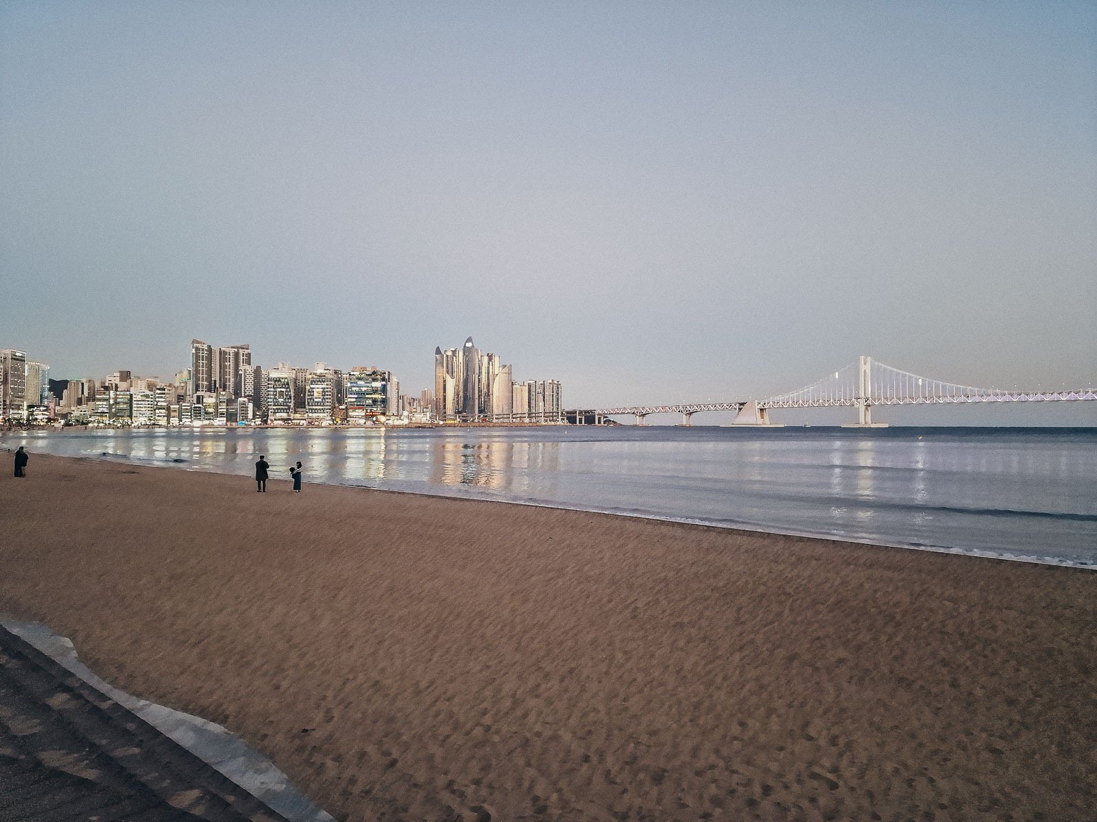 a sandy beach at dusk with a suspension bridge visible in the bay and city skyline lit up on the other side